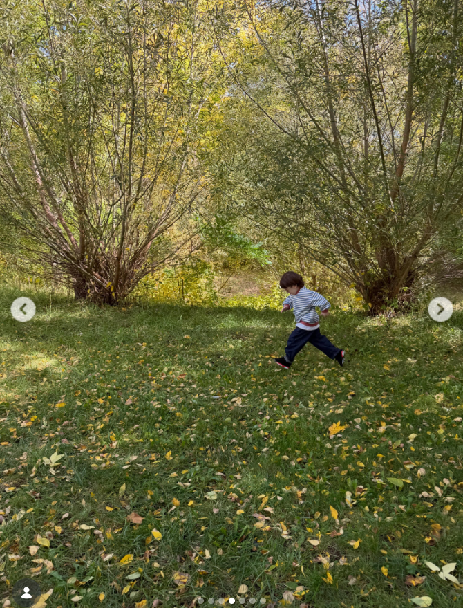 Luai runs across a grassy clearing in a striped shirt, dark pants, and black sneakers with red accents, surrounded by leafy trees and scattered autumn leaves. His dark hair stands out against the golden-green woodland backdrop. | Source: Instagram/lindsaylohan