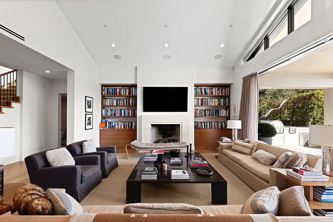 The family room opens directly to a covered outdoor cooking area, extending the gathering space beyond the home's walls. Built-in bookshelves flank the fireplace indoors, while the wide opening puts the backyard within easy reach. | Source: Instagram/tomer.fridman