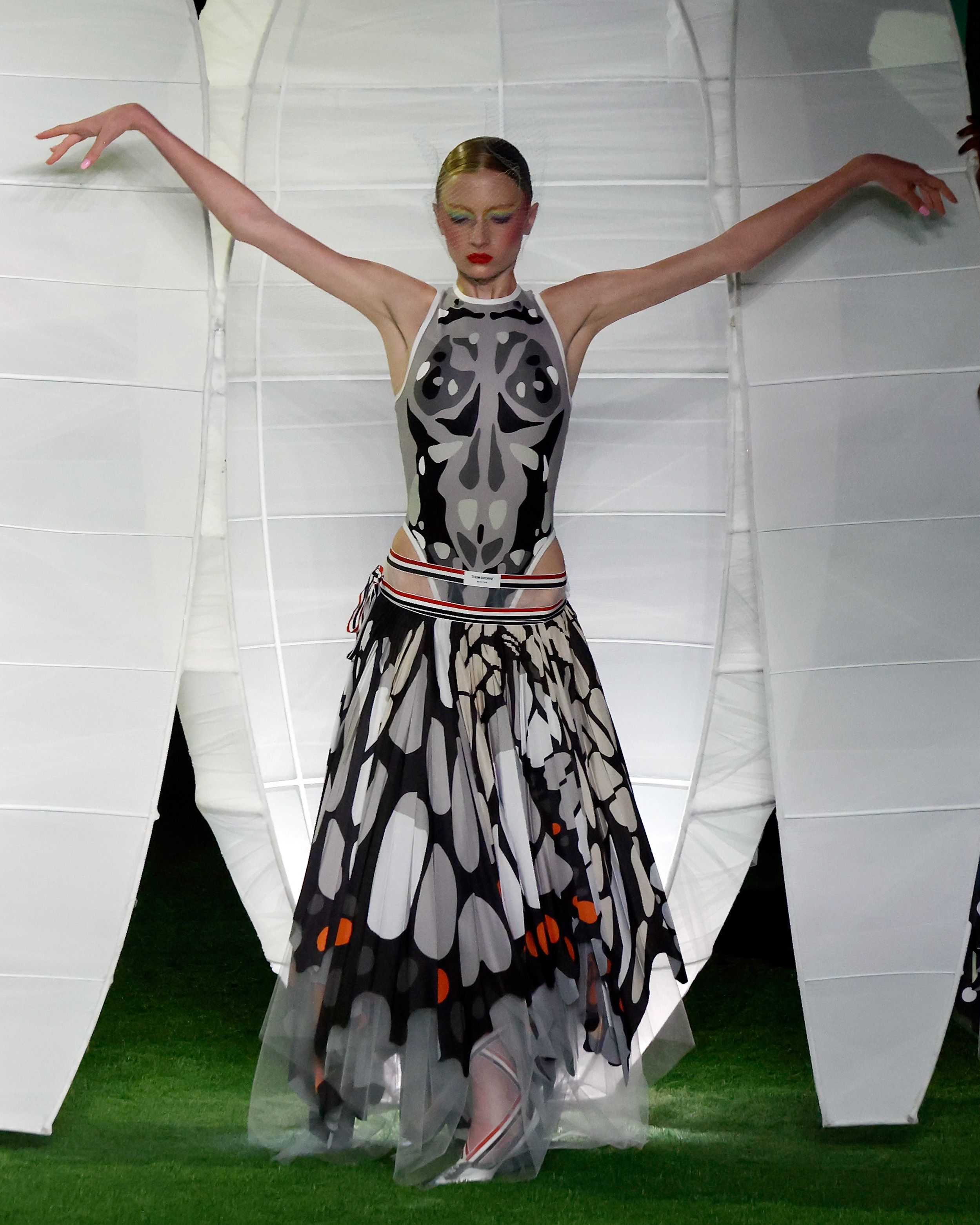 Striking an expressive pose with arms outstretched like wings in front of a giant white cocoon structure, Kai Schreiber embodied theatrical couture at the Thom Browne September 2026 show on September 15, 2026, in New York City. The avant-garde ensemble featured a high-cut illustrated bodice, low-slung striped waistband, and an asymmetrical feathered-pattern skirt complemented by dramatic pastel makeup and a sleek updo. | Source: Getty Images