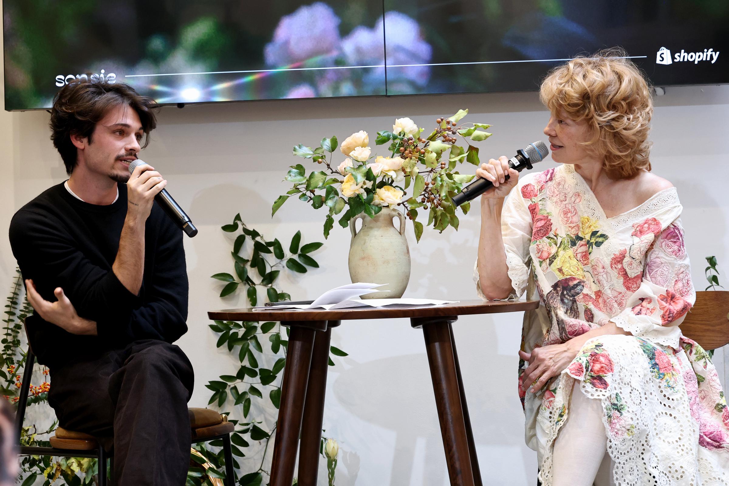 Dylan and Pamela share a lighthearted moment onstage at the "Sonsie Garden" event. Seated beside each other with microphones in hand, they address attendees in front of a floral display and brand signage.