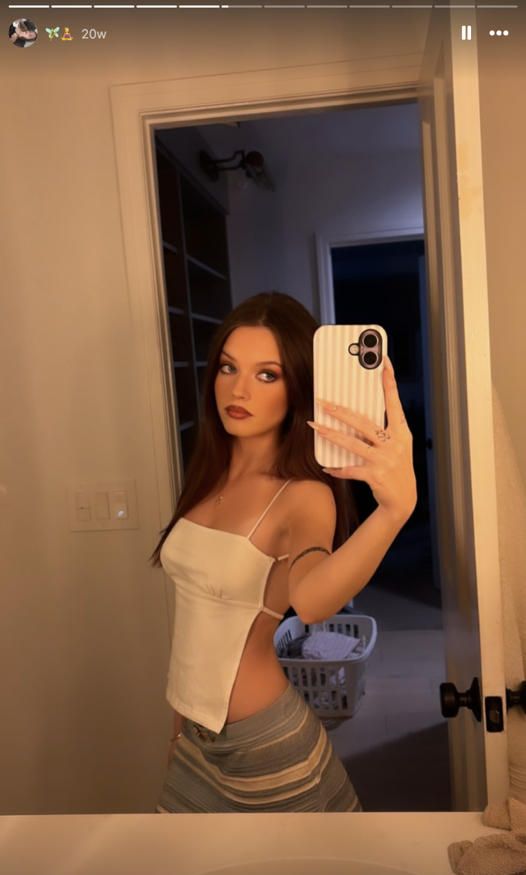Harper Lockwood captures a bathroom mirror selfie wearing an open-back white halter crop top paired with a striped skirt, highlighting her long dark hair and dramatic eye makeup. | Source: Instagram/harper.lockwoodd