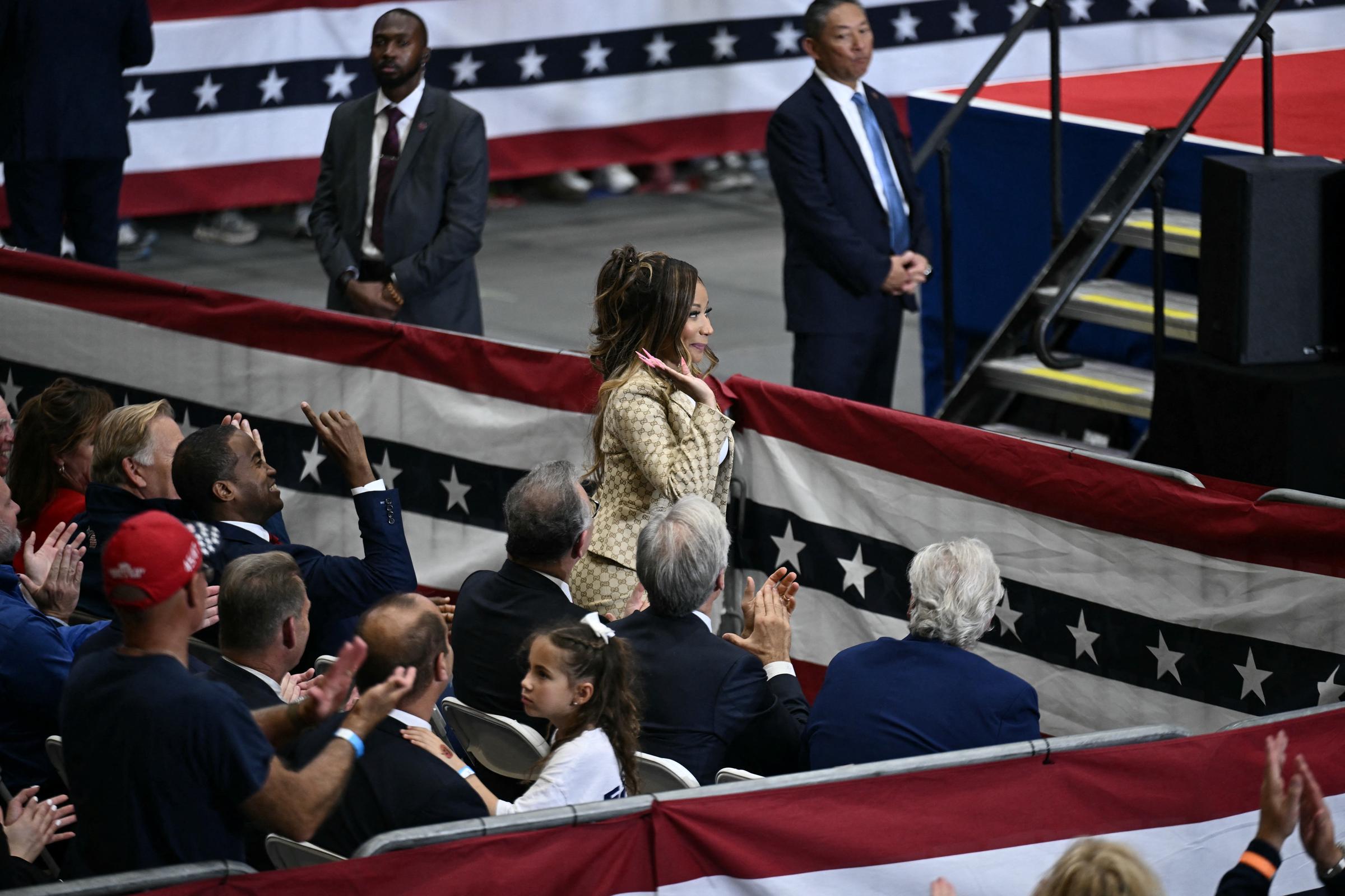 Nicki Minaj waves to applauding attendees at Donald Trump's October 25, 2026, rally in Port Huron, Michigan. The rapper was seated among guests near the stage, wearing a gold-toned Gucci suit that marked a striking departure from her usual performance outfits. | Source: Getty Images