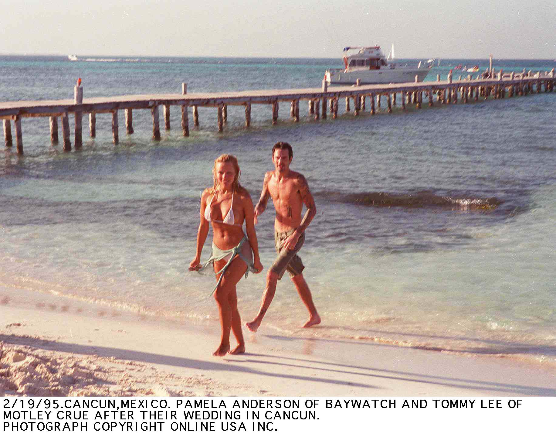 Newlyweds Pamela Anderson and Mötley Crüe drummer Tommy Lee stroll barefoot along the sunny beach in Cancun, Mexico, just days after their whirlwind February 1995 wedding. Wearing a white bikini with a tied sarong alongside Lee in board shorts, the high-profile beach moment marks the intense media storm of Anderson's early fame—a chaotic chapter that would eventually inspire her retreat to the tranquil, grounded solitude of Vancouver Island. | Source: Getty Images