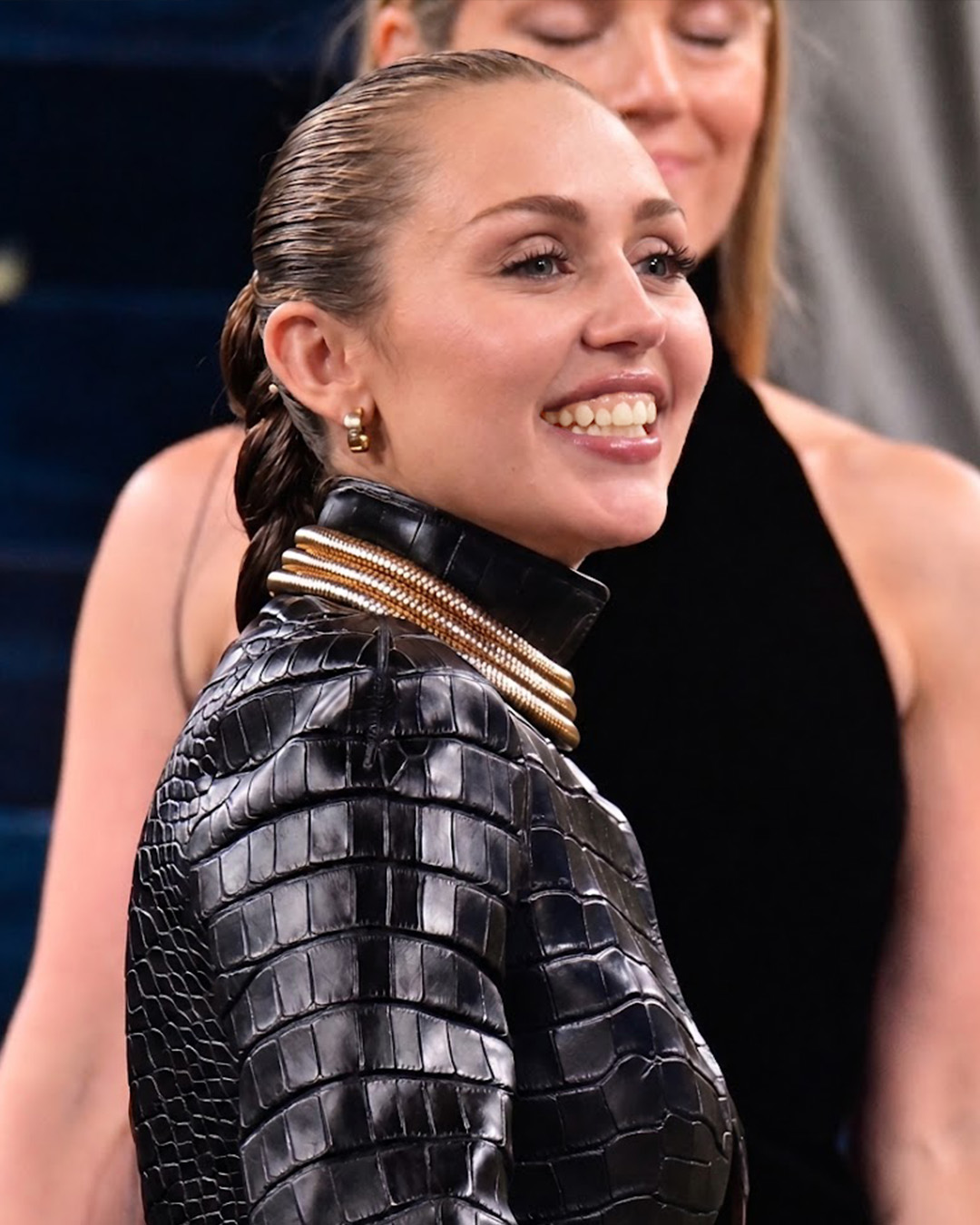 AI-generated image of Miley Cyrus smiling in profile, wearing a glossy black crocodile-textured jacket with a dramatic high collar and layered gold neck jewelry. | Source: Gemini
