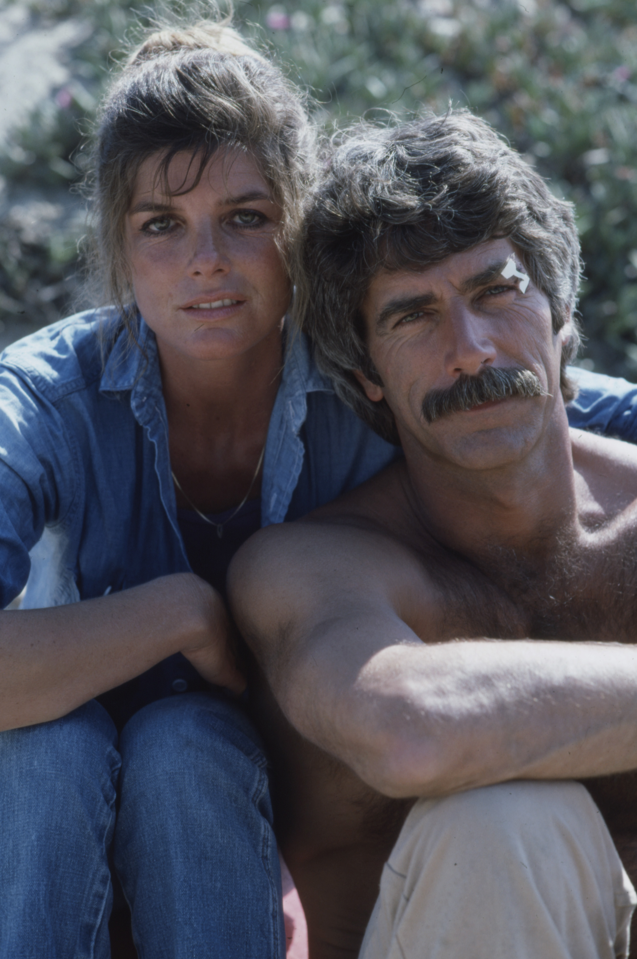 Katharine Ross and Sam Elliott teamed up for a sunlit promotional portrait in Los Angeles, California, in 1983 for the pilot episode of the ABC television series "Travis McGee." Leaning into each other outdoors against lush greenery, Katharine rocked casual double-denim with an open button-down shirt over a dark tee and matching jeans, while a shirtless Sam looked rugged and sun-bronzed with his iconic dark mustache and wavy hair. | Source: Getty Images