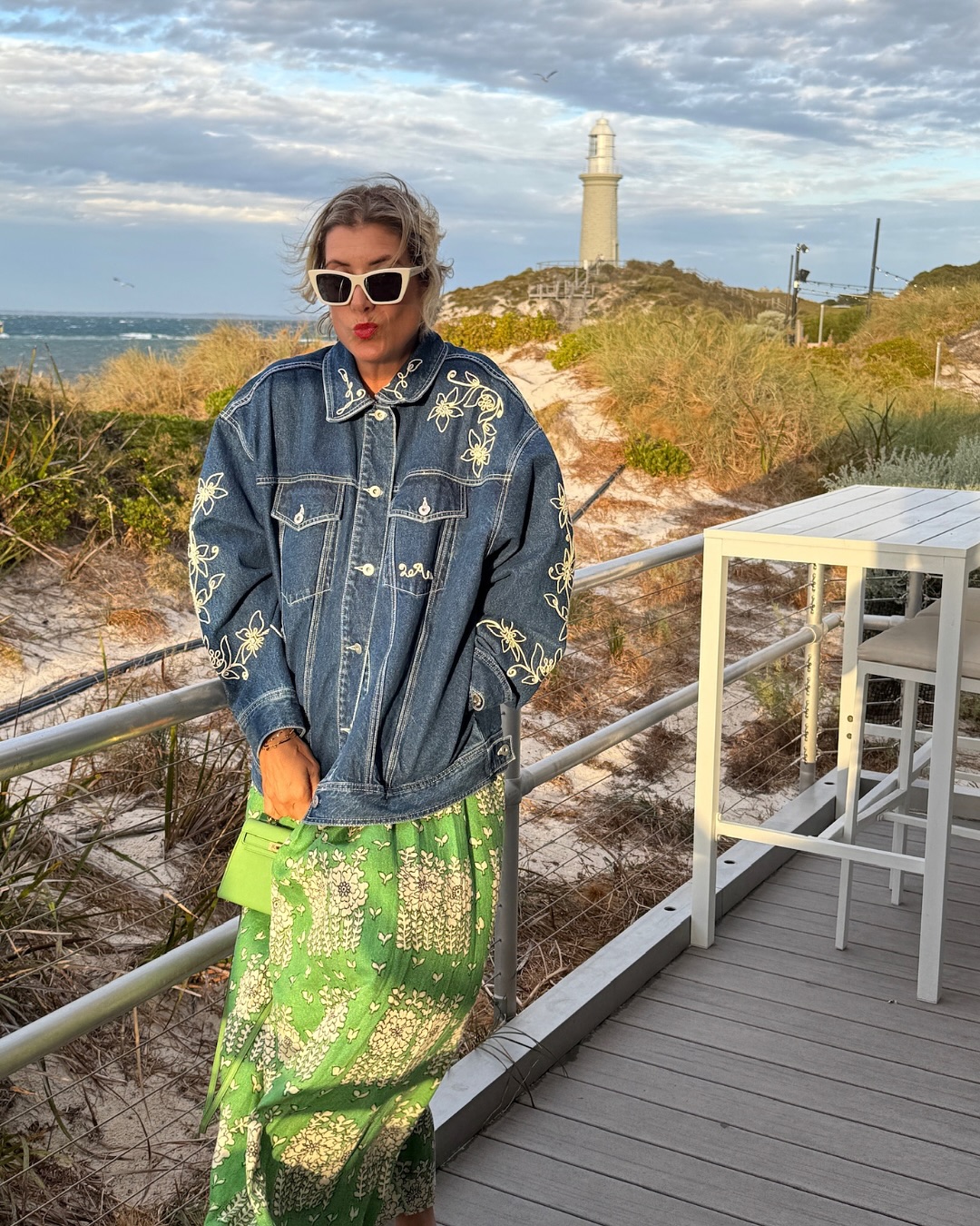 Kate Walsh poses playfully in a floral-embroidered denim jacket and green printed skirt during a seaside outing, dated April 10, 2025 | Source: Instagram/katewalsh