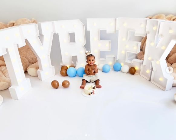 Tyreek Jr. flashes a wide smile while seated in the middle of an elaborate birthday display, complete with oversized illuminated letters spelling "TYREEK," colorful balloons, and large teddy bears. Wearing a tiny crown for the celebratory portrait, Hill and Camille Valmon's son marked his first birthday on March 12, 2024. The image appeared in a post dated March 13, 2024. | Source: Instagram/tyreekhilljr_
