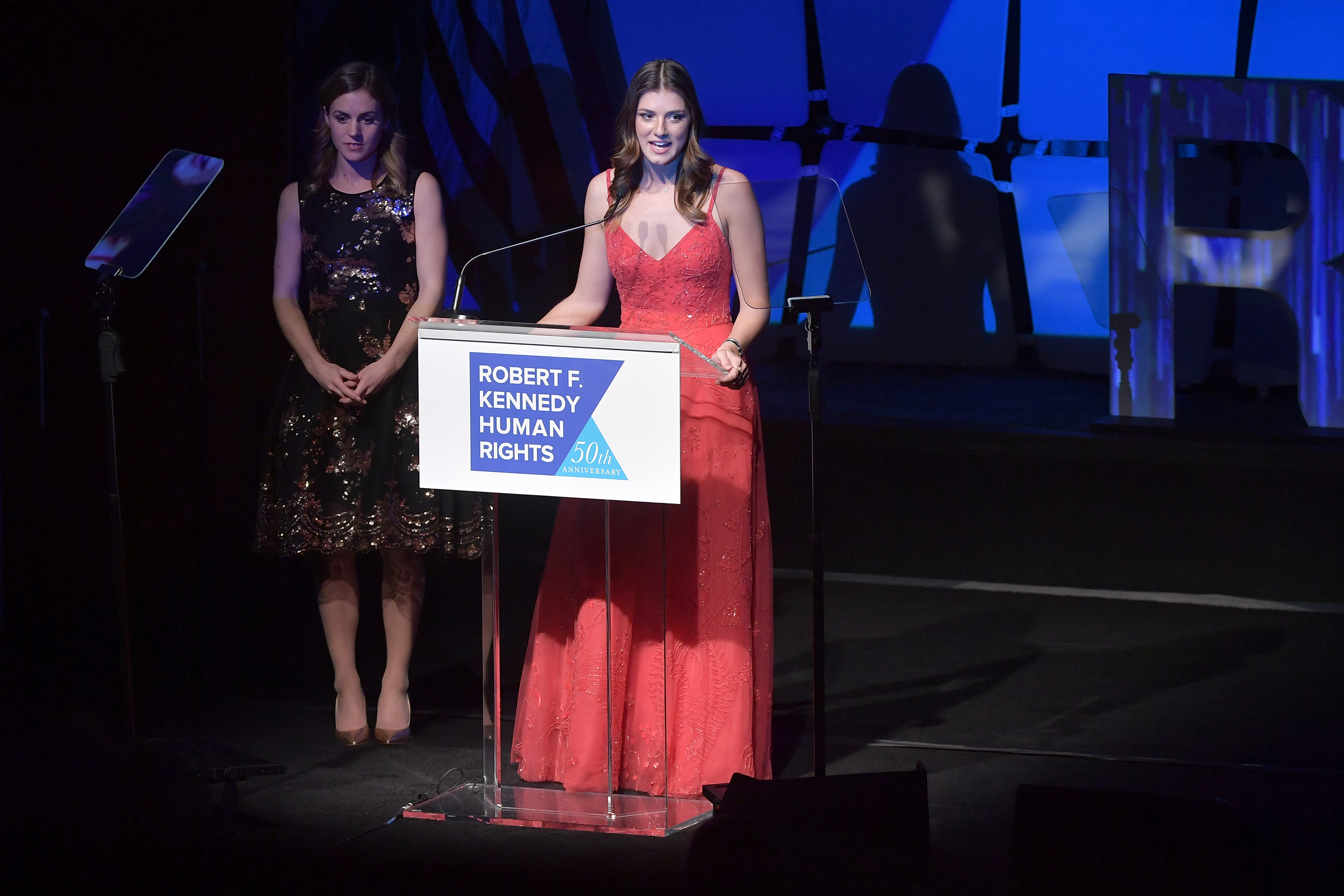 Kyle Kennedy Kerr stands onstage alongside Michaela Kennedy Cuomo during the 2019 Robert F. Kennedy Human Rights Ripple of Hope Awards on December 12, 2018, in New York City. Michaela addresses the crowd from behind the clear podium in a coral-red spaghetti-strap evening gown, while Kyle looks on attentively in a sleeveless knee-length black dress embellished with shimmering bronze sequins and metallic heels. | Source: Getty Images