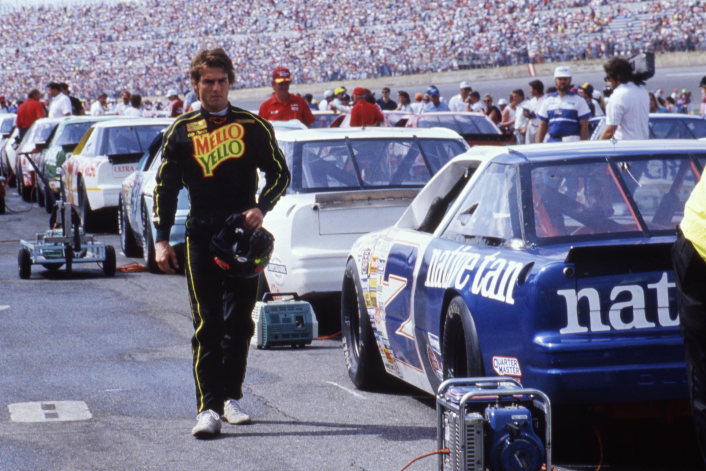 Tom Cruise appears as Cole Trickle on the set of "Days of Thunder," walking through a crowded track area in his black racing suit while carrying his helmet. Stock cars and crew members fill the background of the racing scene from the 1990 film. | Source: Getty Images