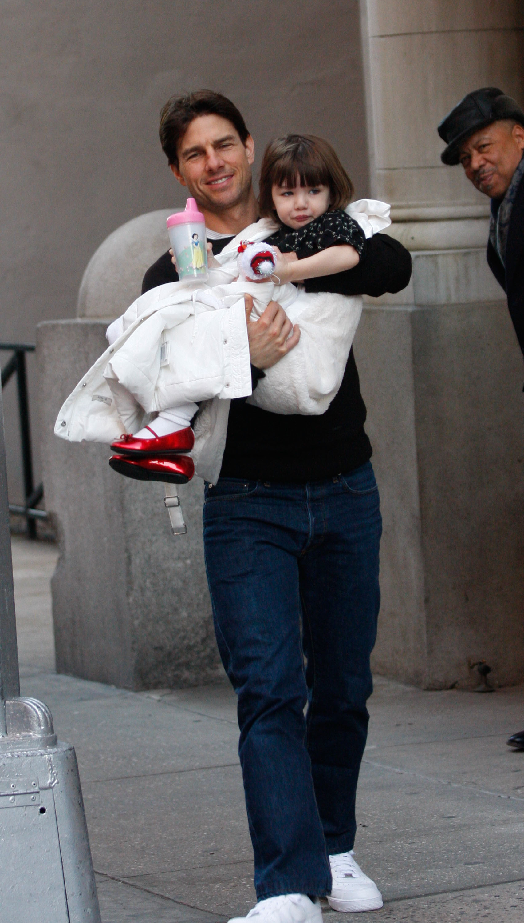 Captured in a tender street-side moment, Tom Cruise carries baby Suri in his arms as cameras snap away. With a bottle of milk in hand and her gaze wandering, Suri appears calm and curious amid the bustle. Tom, focused and composed, shields his daughter with a protective hold.