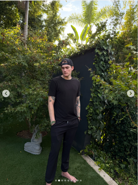 Presley Gerber turns slightly toward the camera with a subtle smile, standing barefoot in the garden with his tattoos visible along his arms and one hand tucked into his pocket. | Source: Instagram/presleygerber