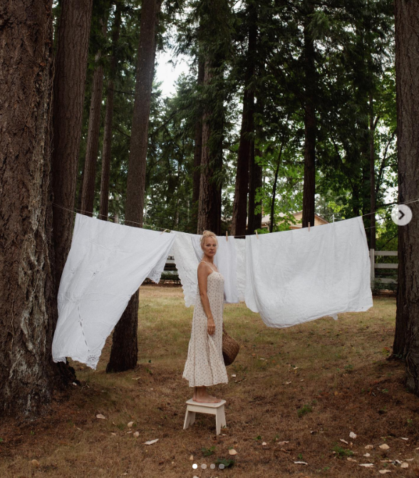 Standing barefoot atop a white wooden step stool, Pamela Anderson pauses while hanging laundry between soaring evergreens at Arcady. Wearing a delicate floral sundress with her hair swept up, she gazes into the camera framed by fluttering white linens, captioning the pastoral ritual: "I look forward to the morning chores… I feed and walk the dogs, I do the laundry, I write… I feel my life wrapped humbly around me. All is well 🤍". | Source: Instagram/pamelaanderson