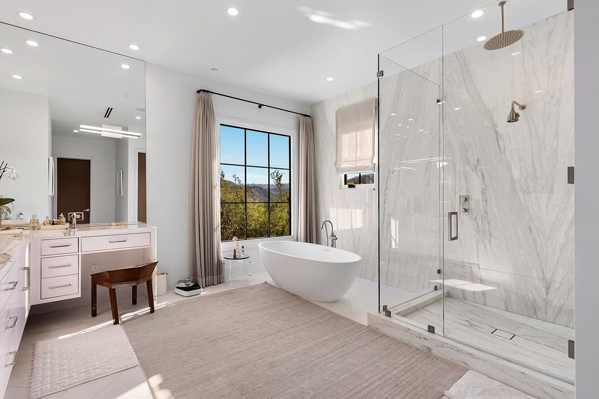 The primary bathroom returns to the home's lighter aesthetic with white marble, a glass-enclosed shower, and a freestanding soaking tub. A large window beside the tub brings in natural light and opens the room to views of the surrounding hills. | Source: Instagram/tomer.fridman