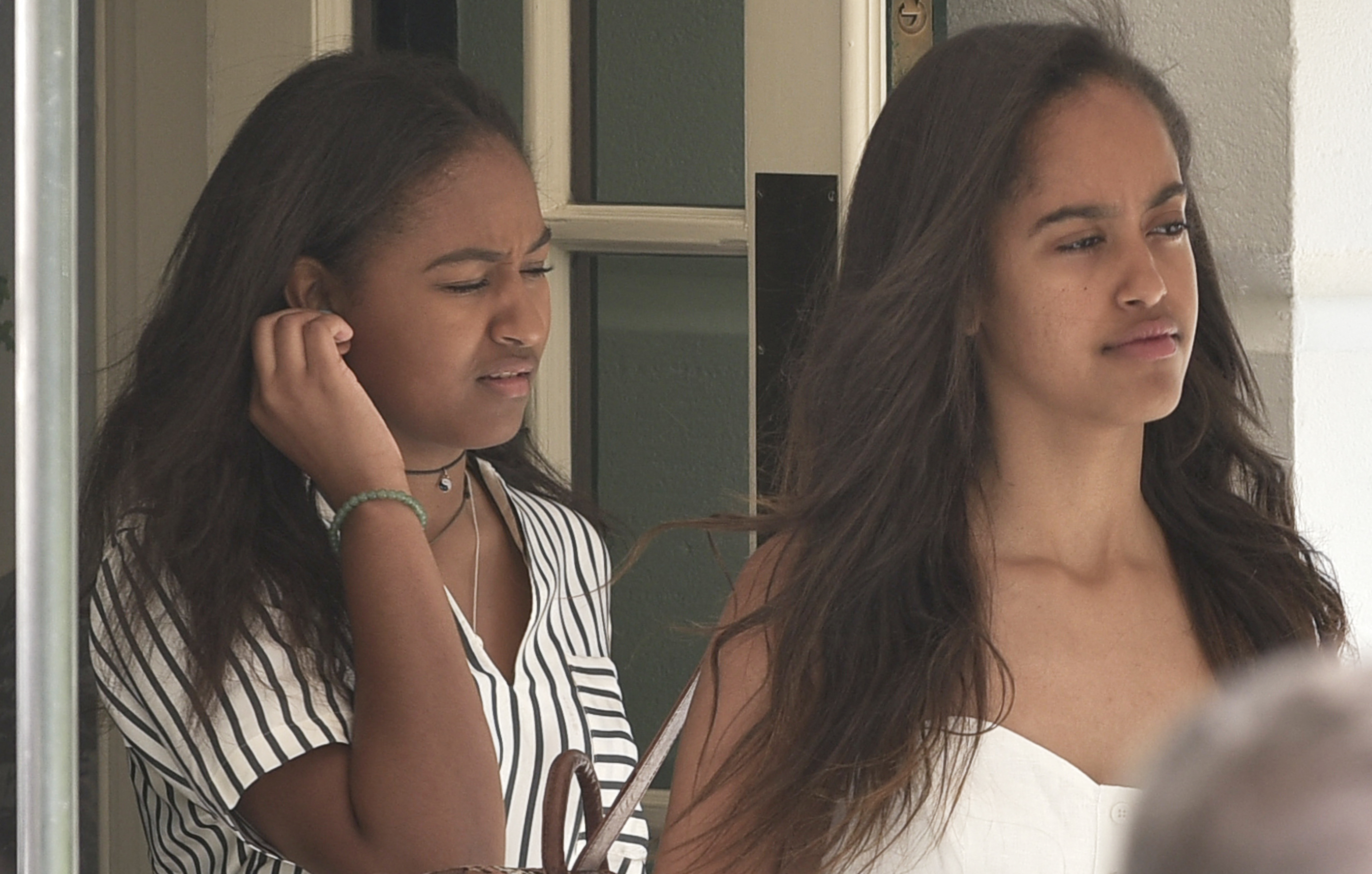 Ahead of their summer flight on August 6, 2016, in Washington, D.C., Sasha Obama, in a striped blouse and black choker, squints in the sun beside Malia. Her sister, wearing a white sundress, looks ahead with a calm and observant expression.