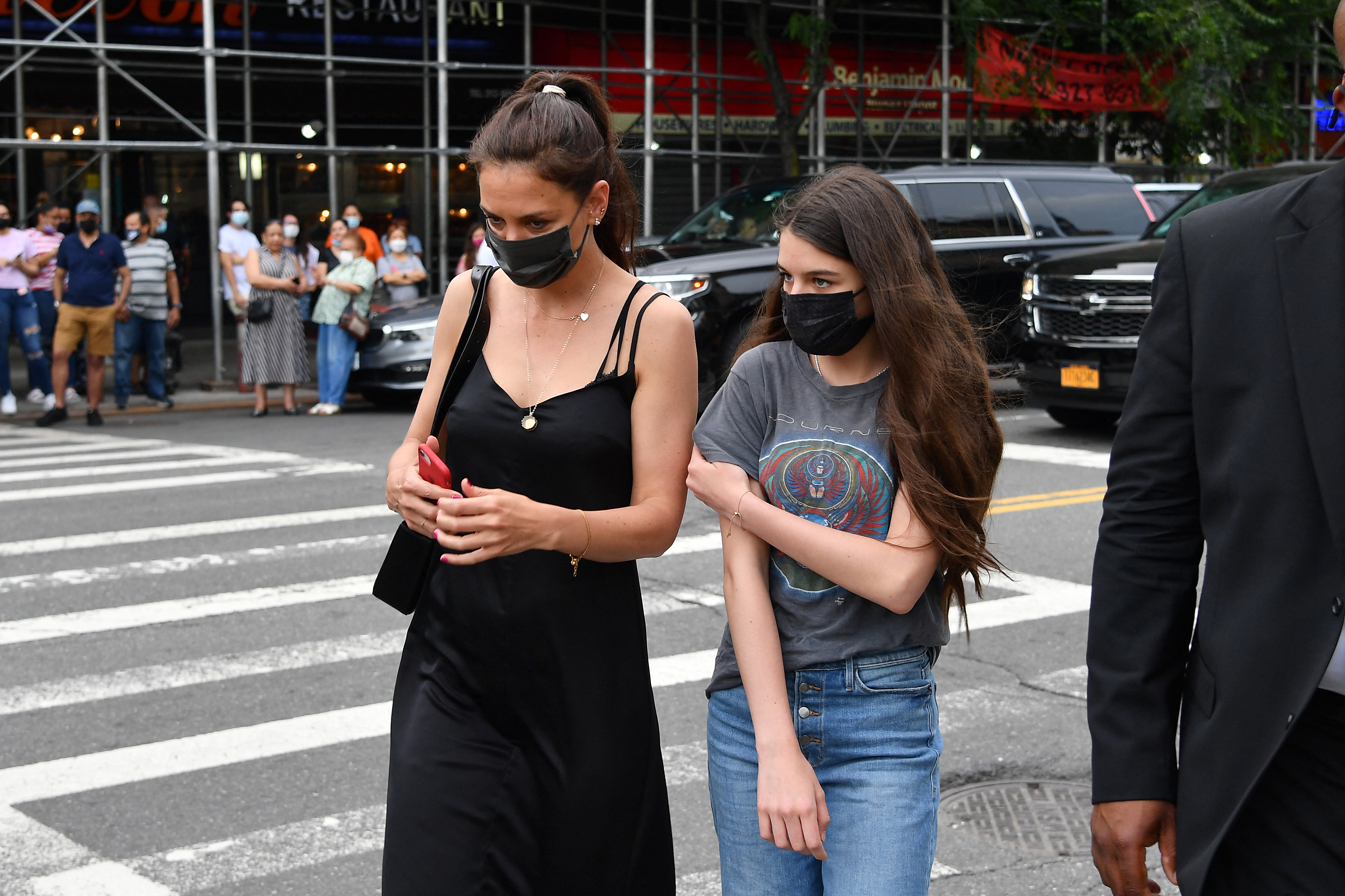 Suri Cruise and her mother, Katie Holmes, are captured mid-stride as they cross a city street, both wearing black face masks. The coordinated look adds a sleek touch to their casual outing, while their focused expressions reflect the quiet caution of pandemic life.