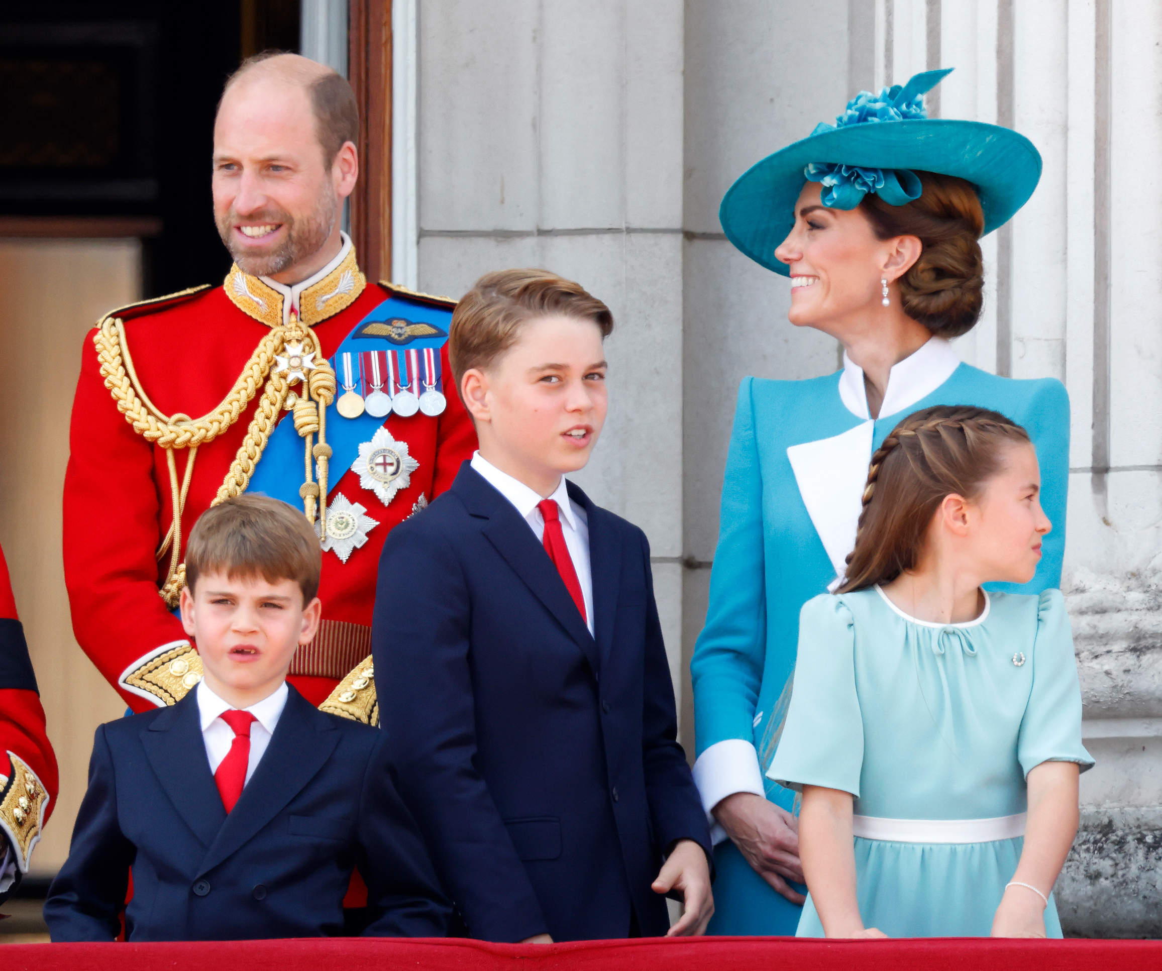 Princess Charlotte, Princess Catherine, Prince William, Prince Louis, and Prince George stand on the balcony of Buckingham Palace. Prince William and Princess Catherine smile while Princess Charlotte looks sideways, giving a beautiful view of her sleek hairdo that has social media buzzing.