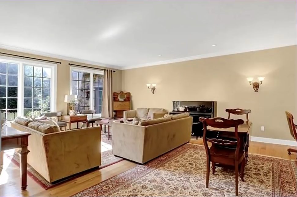 This spacious living and dining area features beige walls, hardwood flooring, traditional wooden furniture, and patterned rugs. Large windows brighten the sitting area, while a fireplace provides a focal point along the far wall. | Source: YouTube/Without A Crystal Ball