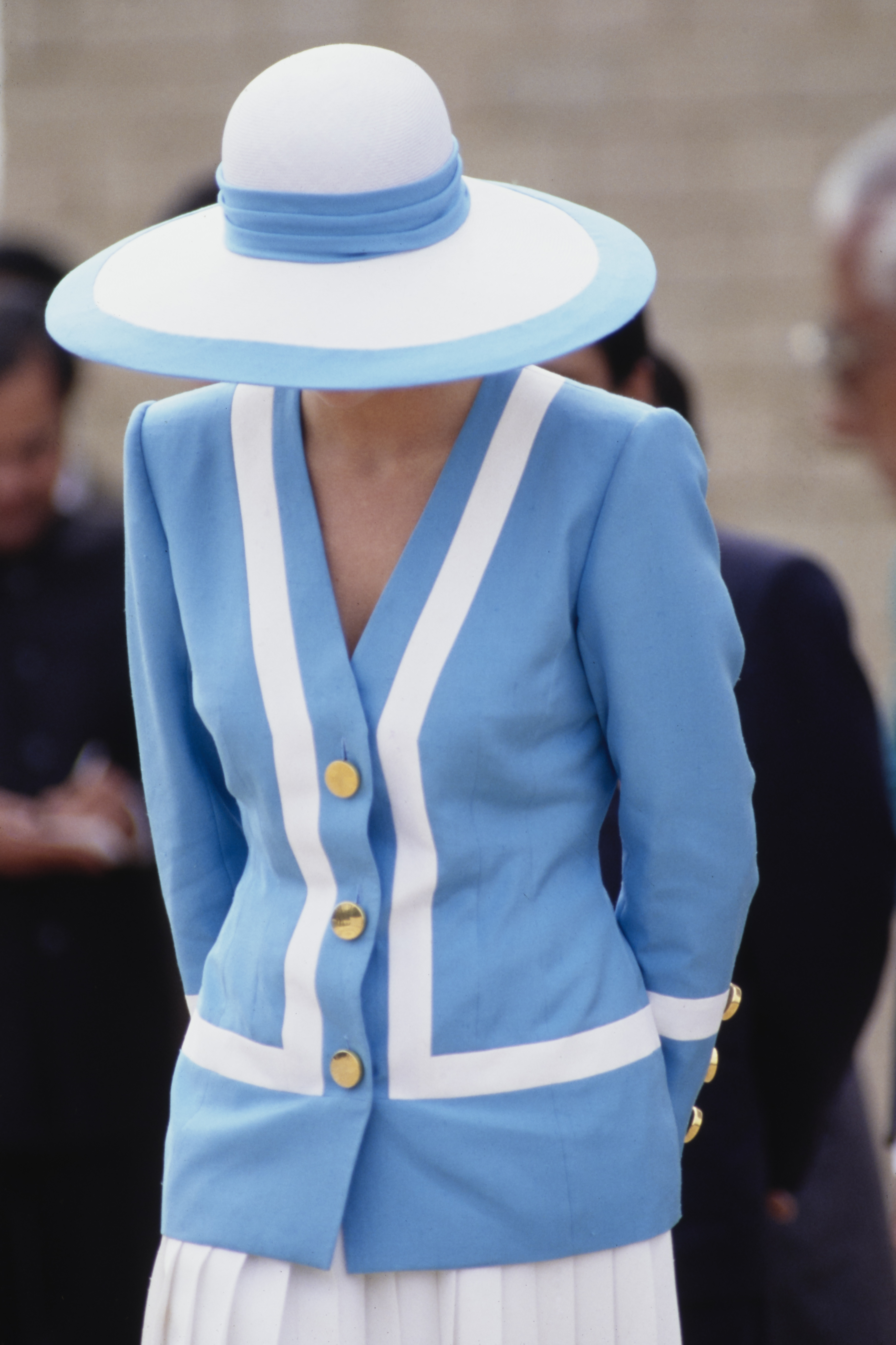 Princess Diana stands with quiet poise at the Delhi War Cemetery, dressed in her signature blue and white suit and a wide-brimmed hat. The image captures her solemn grace amid the tranquil setting, a reflection of respectful remembrance.