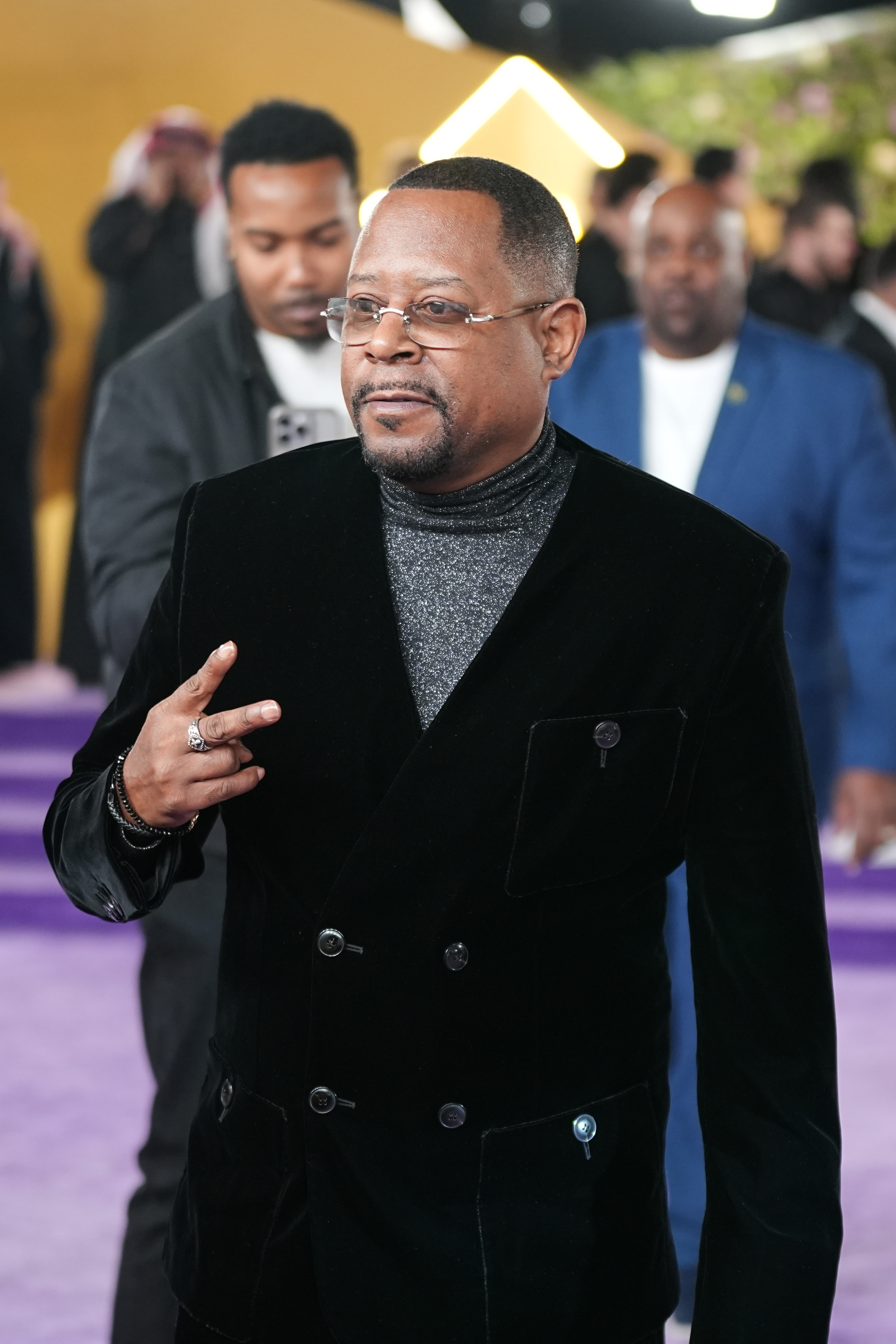 At the 2025 Joy Awards in Riyadh, Martin Lawrence wears a black velvet suit layered over a shimmering turtleneck. He flashes a peace sign while walking through the venue, blending elegance with his signature laid-back style.