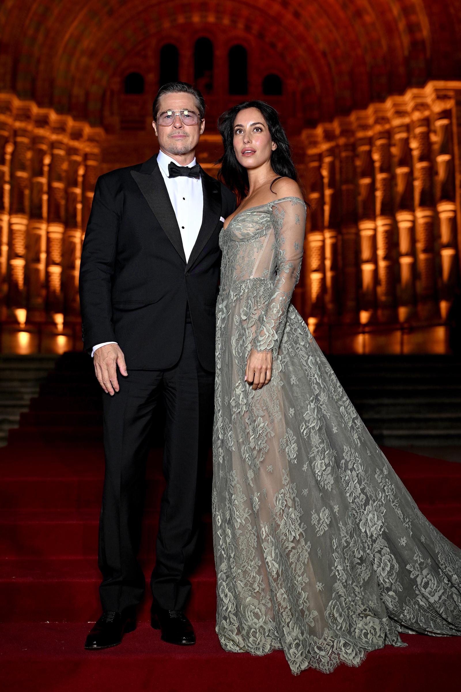 Brad Pitt and Ines de Ramon pose for a full-length photograph on the red-carpet staircase, showcasing their contrasting formal looks. Ines's floor-length embroidered gown features a dramatic train that fans out across the steps, while the venue's glowing golden arches create a striking backdrop. | Source: Getty Images