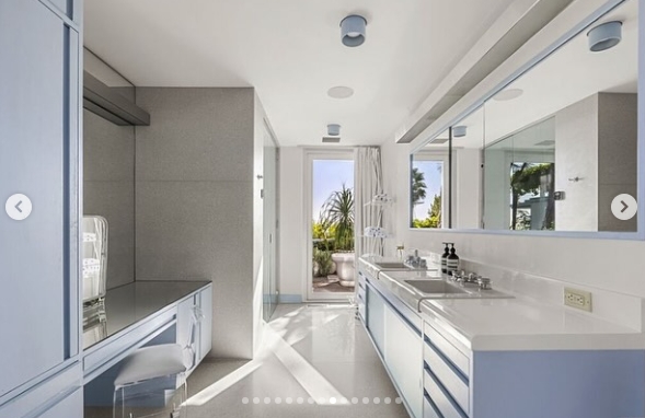 The primary bathroom continues the home's crisp aesthetic with long countertops, pale blue cabinetry, expansive mirrors, and a streamlined layout. Sunlight enters from the doorway leading outside, where greenery is visible beyond, reinforcing the property's recurring blend of private interior spaces and open-air living. | Source: Instagram/jparkerstanton