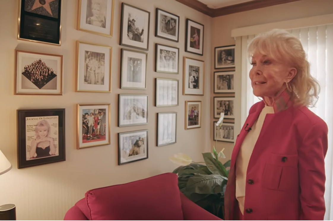 Barbara Eden stands beside a red upholstered armchair in her home, surrounded by a gallery wall of framed photographs, magazine covers, and memorabilia documenting different moments from her life and career. | Source: YouTube/People