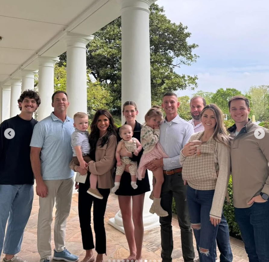 Bryon and Kristi Noem pose with family members along a column-lined outdoor walkway, smiling together as several adults hold young children. | Source: Instagram/kristinoem