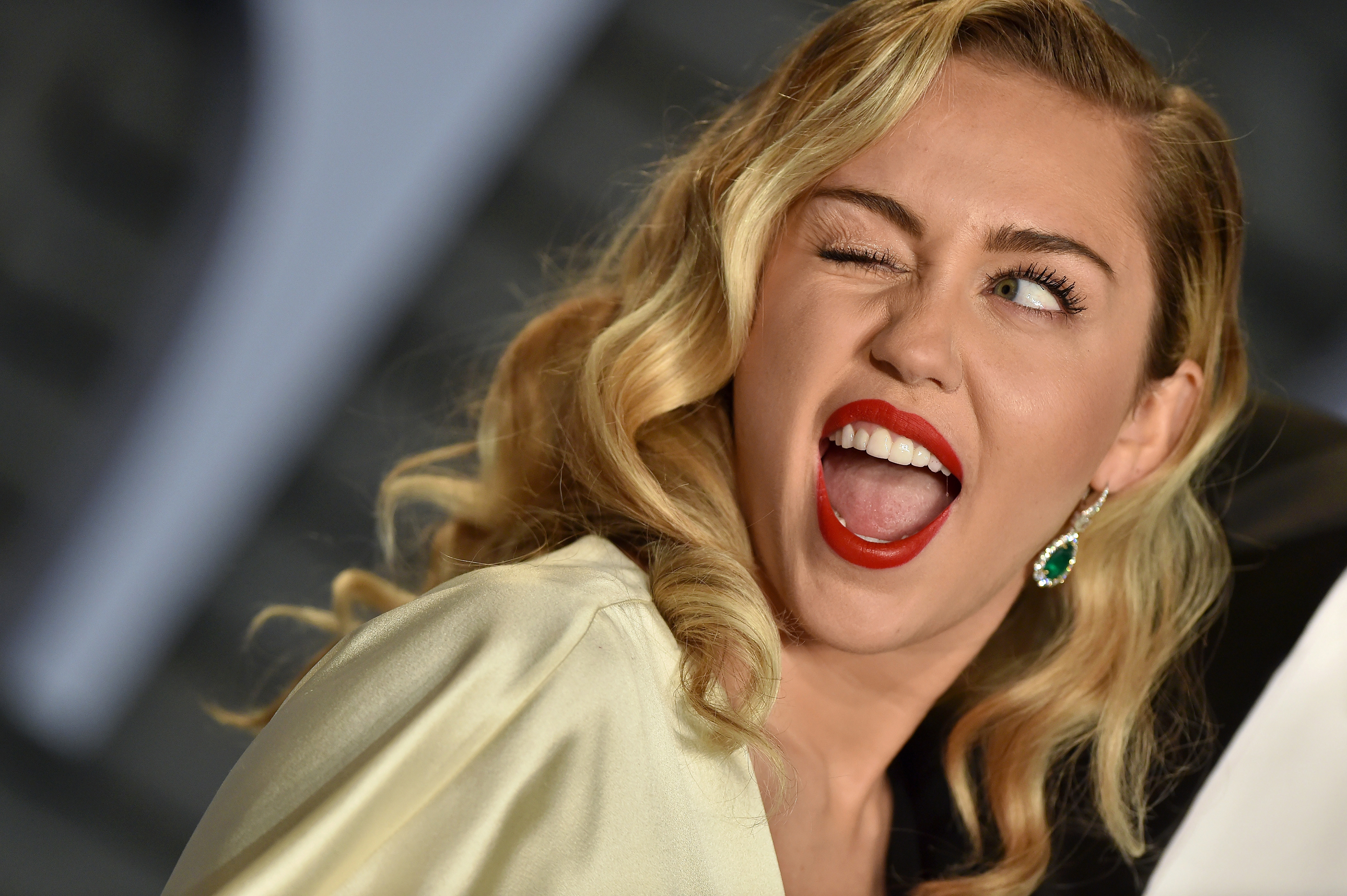 Miley Cyrus struck a playful pose with one eye closed, wearing bold red lipstick, emerald earrings, and a pale gold outfit with her blonde hair styled in loose waves. Her expressive smile offers a different view of the feature Lange discussed when speculating about changes to Cyrus’s upper lip and the visibility of her gums. | Source: Getty Images