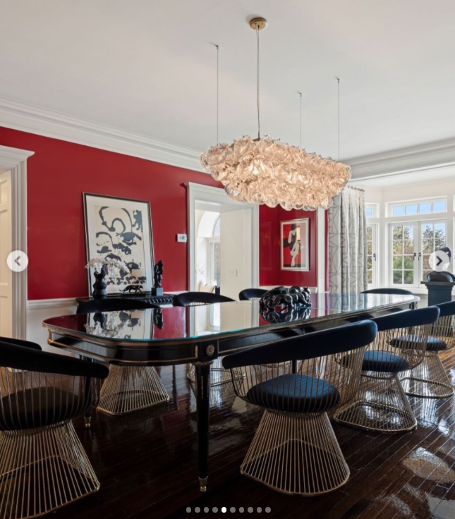 Red walls make the dining room one of Longmeadow's boldest spaces, while a large chandelier hangs above the glossy table. Modern artwork and wire-frame chairs give the formal room a contemporary edge. | Source: Instagram/rogerperrygroup