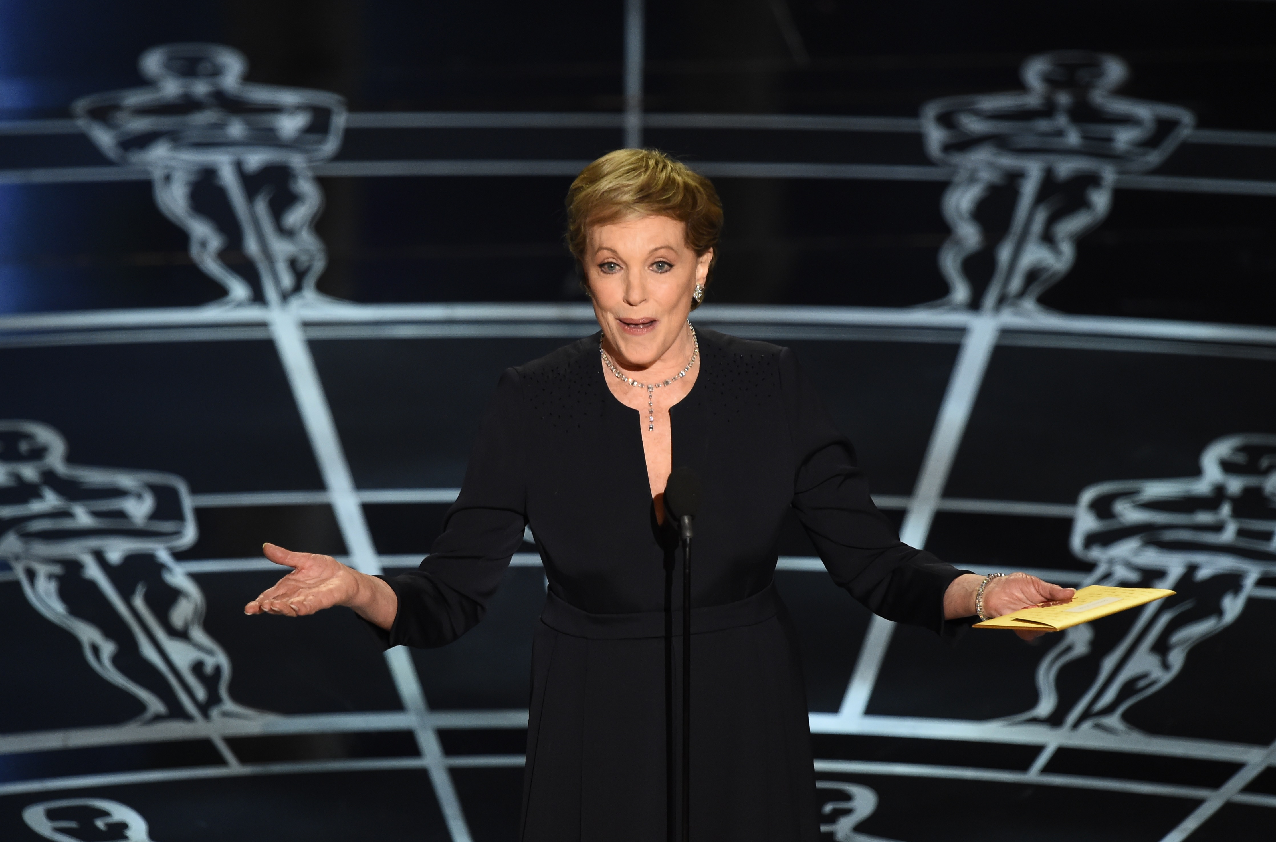 Julie Andrews addresses the audience on stage with open arms at the 87th Academy Awards at the Dolby Theatre in Hollywood, California, on February 22, 2015. Appearing after Lady Gaga's 50th-anniversary musical tribute to "The Sound of Music" to present the Oscar for Best Original Score, Andrews looks timelessly chic holding the gold envelope in an elegant black long-sleeved gown with subtle shoulder embellishments, complemented by an intricate diamond necklace and matching earrings.