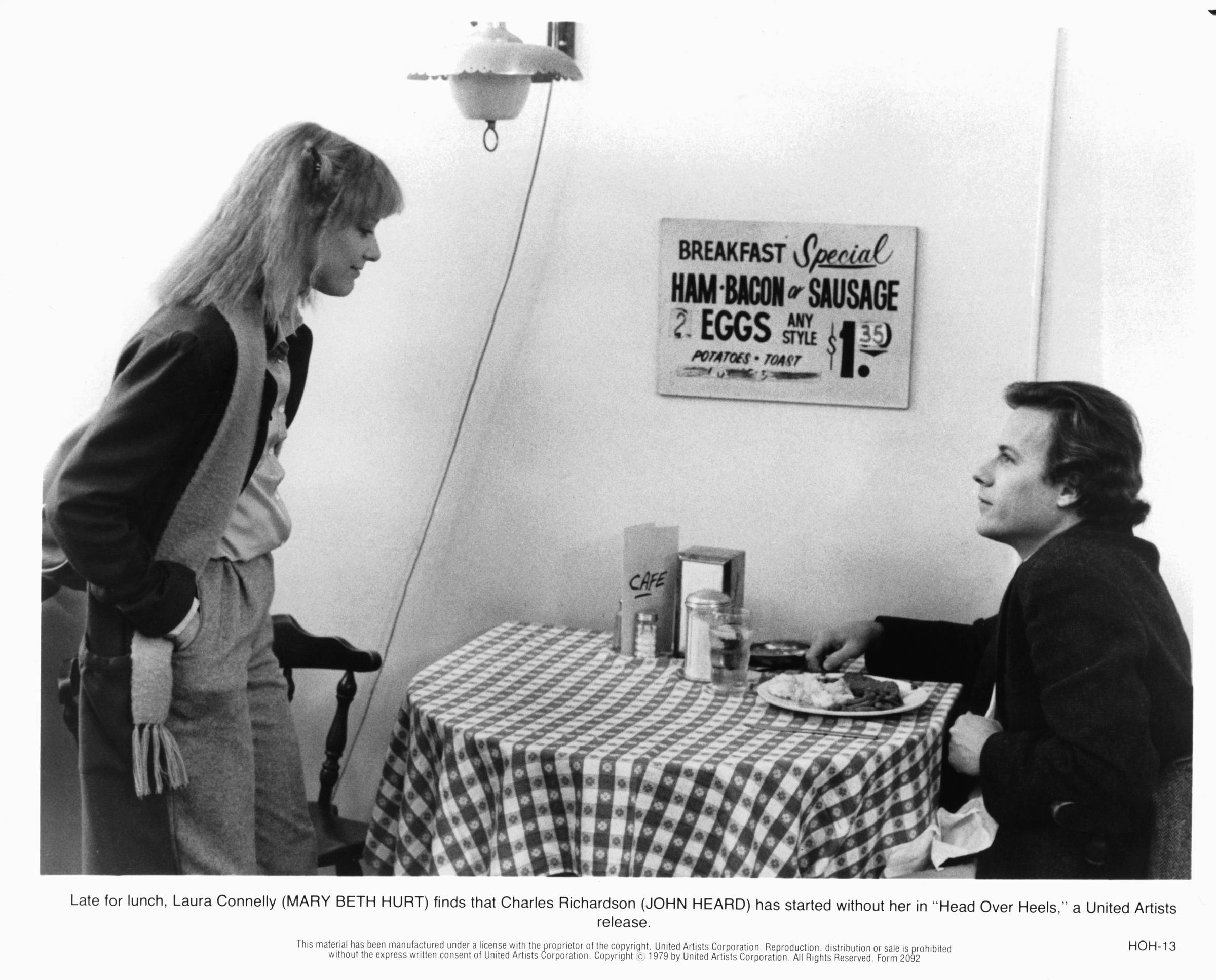 Mary Beth Hurt faces John Heard across a small diner table in "Head Over Heels" in 1979, wearing a casual layered outfit. The scene captures a tense yet playful moment as Hurt's character realizes Heard's character has started lunch without her.
