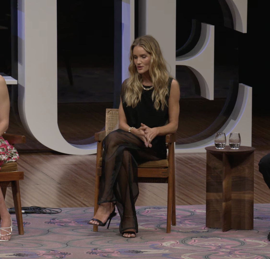 Rosie Huntington-Whiteley sits poised on stage during a panel discussion, dressed in a flowing black ensemble with sheer layers and strappy heels. | Source: YouTube/VogueAustralia