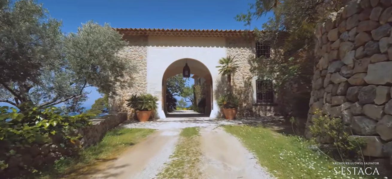 Stone walls and a wide arch give this entrance a warm Mediterranean feel. Potted palms frame the passage, while the blue water visible in the distance is a reminder of the home's coastal setting. | Source: YouTube/billionsluxuryportal