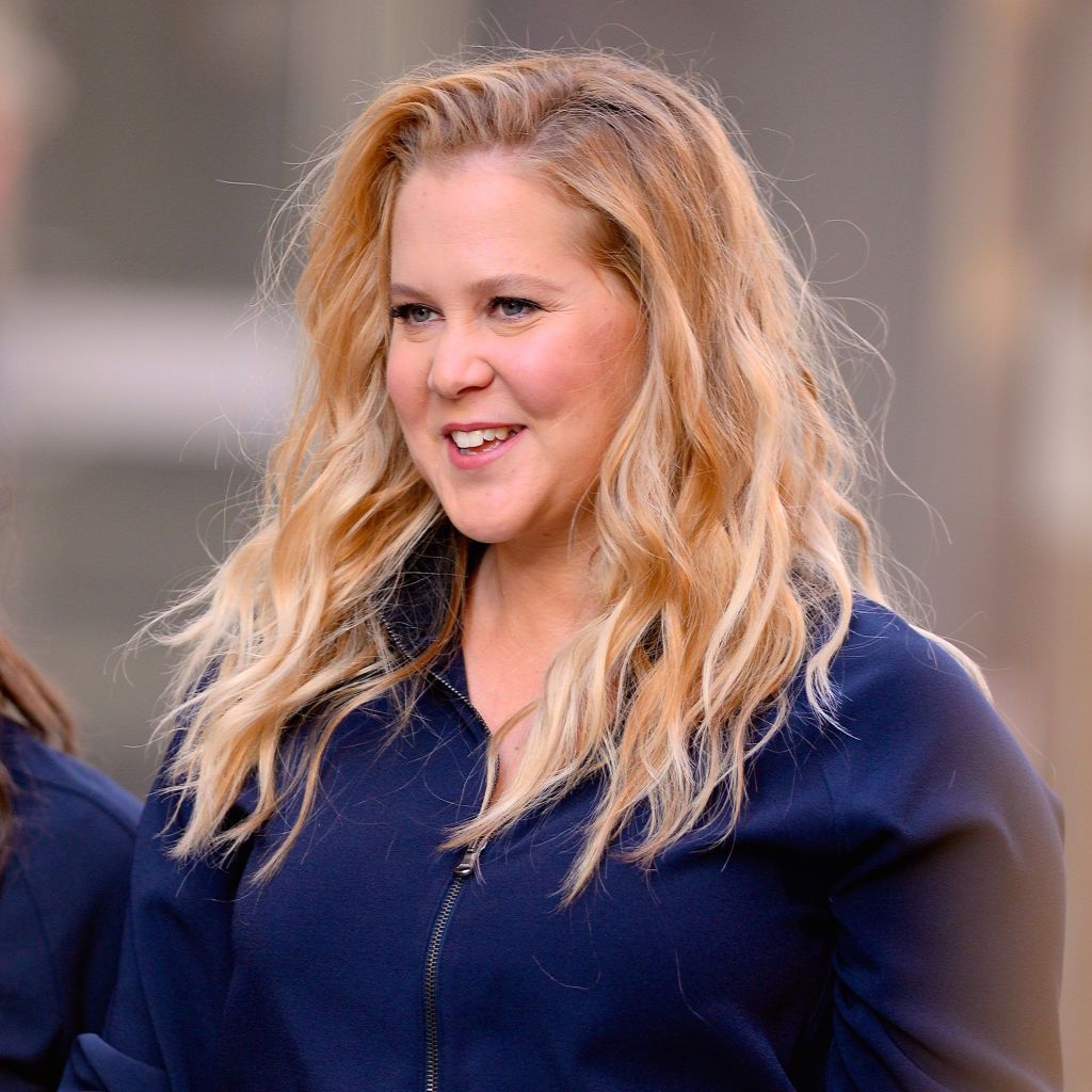 Amy Schumer is seen smiling while filming a commercial on the streets of SoHo in New York City. She wears a navy zip-up top with her wavy blonde hair loose around her shoulders.