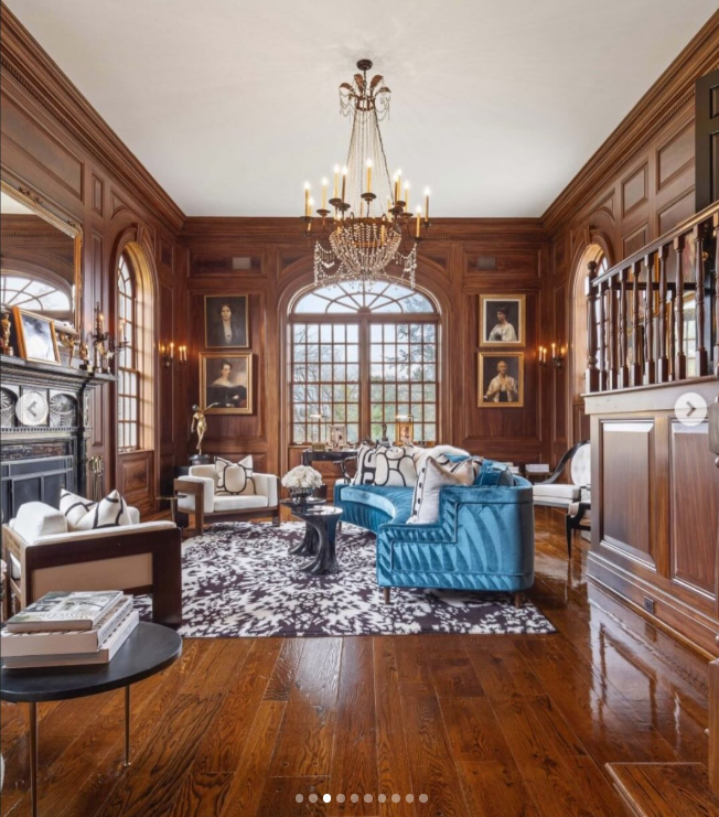 Dark wood paneling wraps this soaring room, where arched windows, portraits, a chandelier, and polished floors create a distinctly traditional atmosphere. Bright blue seating brings a bold burst of color to the otherwise warm, wood-heavy space. | Source: Instagram/rogerperrygroup