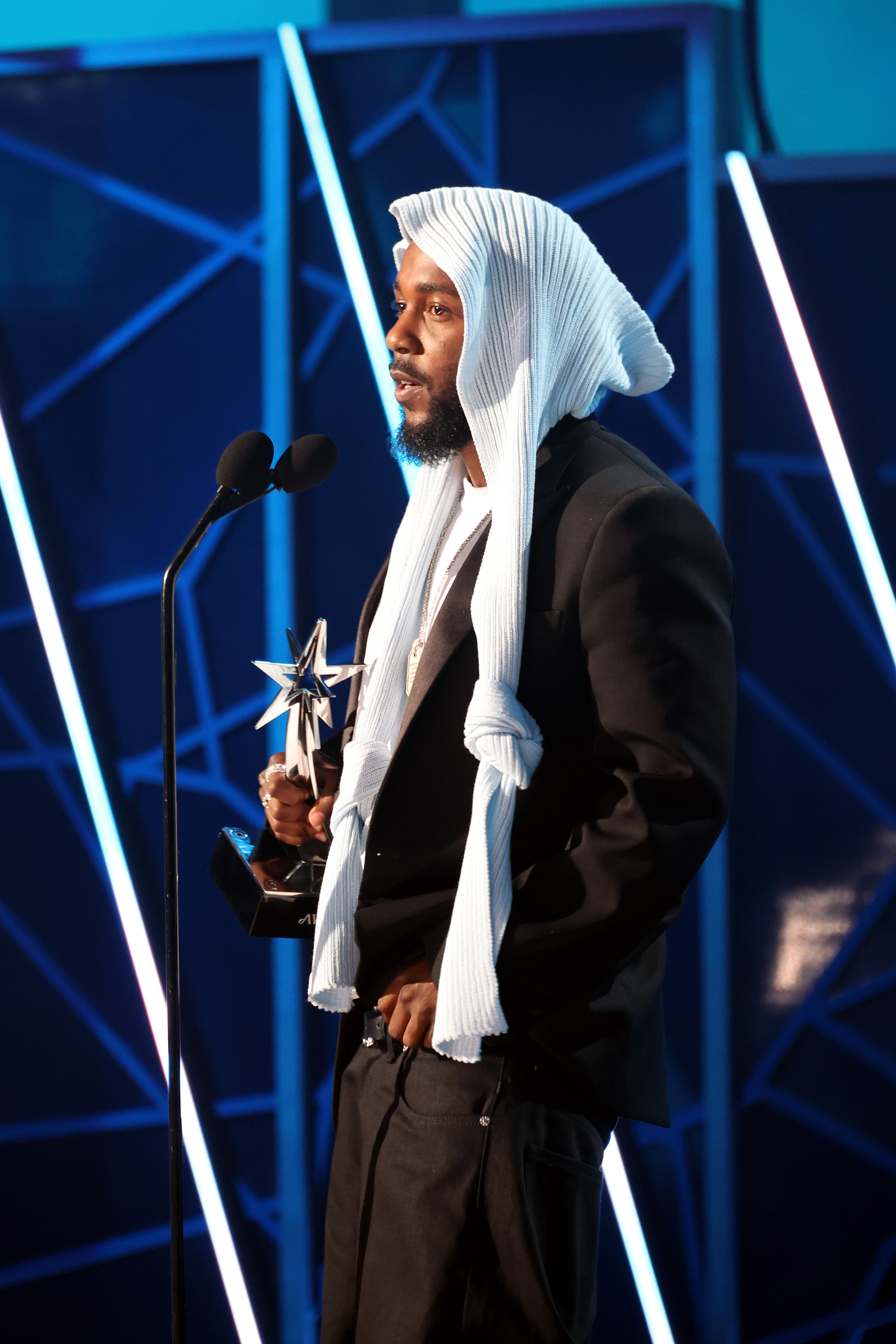 With a calm yet powerful presence, Kendrick Lamar addresses the audience after winning at the 2025 BET Awards. Dressed in a black-and-white ensemble, the artist held his trophy proudly as he stood before the glowing geometric backdrop.
