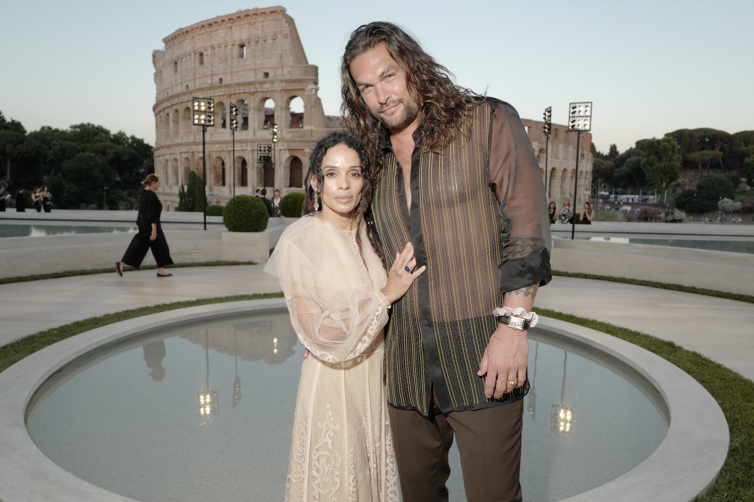 Jason Momoa and Lisa Bonet arrive at the Shine On Sierra Leone 5th Annual Fundraiser on May 25, 2011, in Venice, California. Momoa embraces Bonet while sporting shoulder-length wavy hair, a distressed leather motorcycle jacket, dark trousers, and a black dotted scarf draped over a dark tee, complementing Bonet's layered bohemian style featuring dreadlocks, an ornate coin necklace, and a dark vest over a flowing tunic. | Source: Getty Images