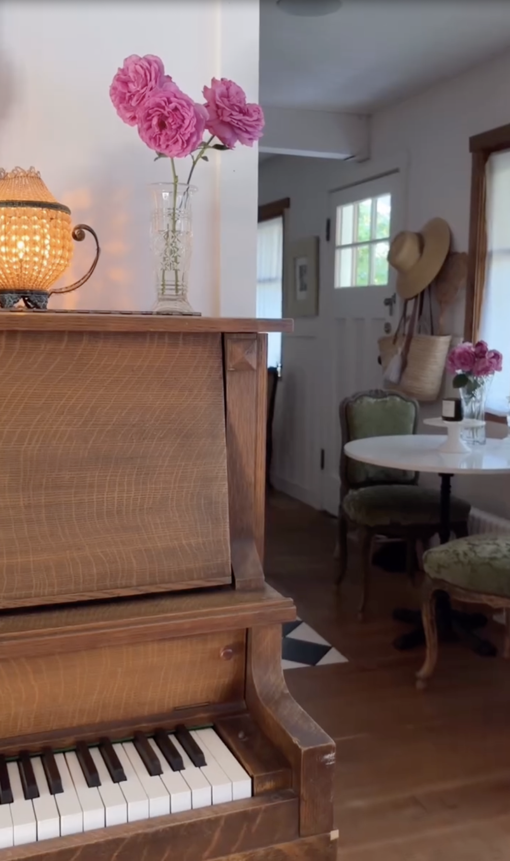A sunlit corner of Pamela Anderson's Arcady retreat highlights her cozy, musical life, featuring an upright wooden piano alongside freshly clipped pink garden roses displayed in cut-crystal glassware. In the background, straw sun hats hang near the door beside a cafe table and green velvet chairs, capturing her serene, self-made romance. | Source: Instagram/pamelaanderson