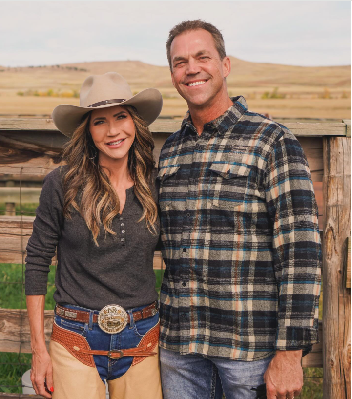 Bryon Noem stands beside Kristi Noem outdoors, both smiling at the camera in a rural setting with open fields and wooden fencing in the background. | Source: Instagram/sdbryonnoem