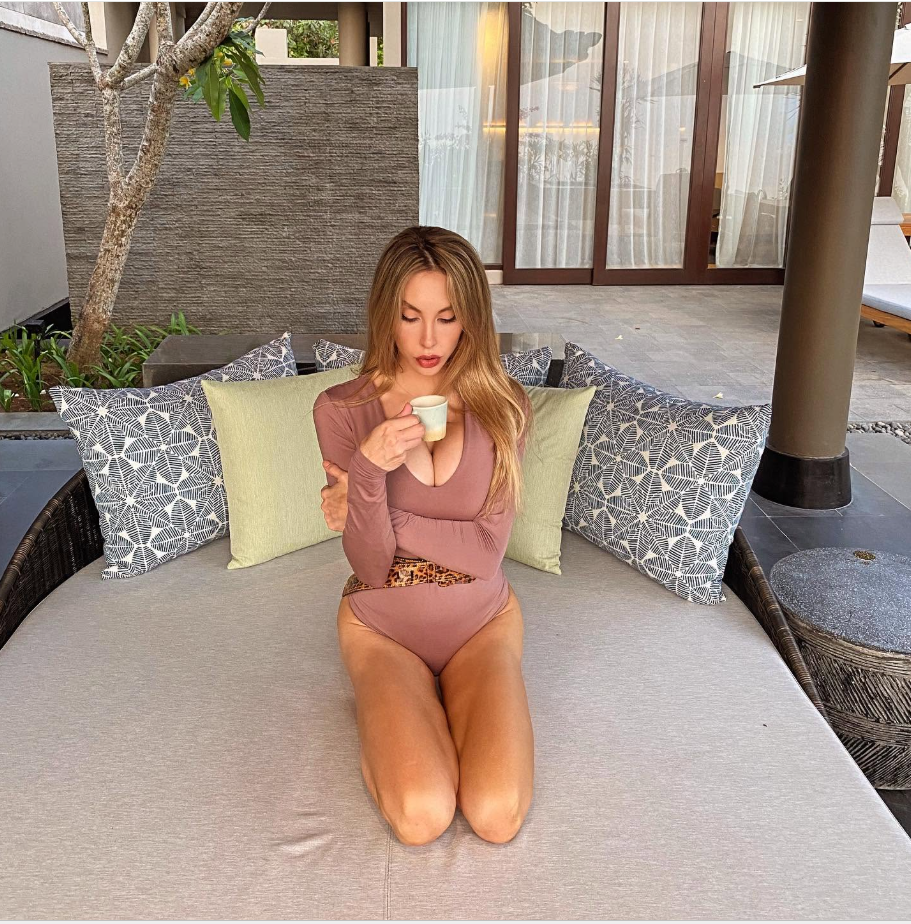 Chloe Lattanzi sits on an outdoor daybed at a tropical-style residence, holding a small cup while wearing a mauve long-sleeved swimsuit with a leopard-print accent. Patterned cushions, a stone wall, and sliding glass doors surround the seating area. | Source: Facebook/Chloe Lattanzi