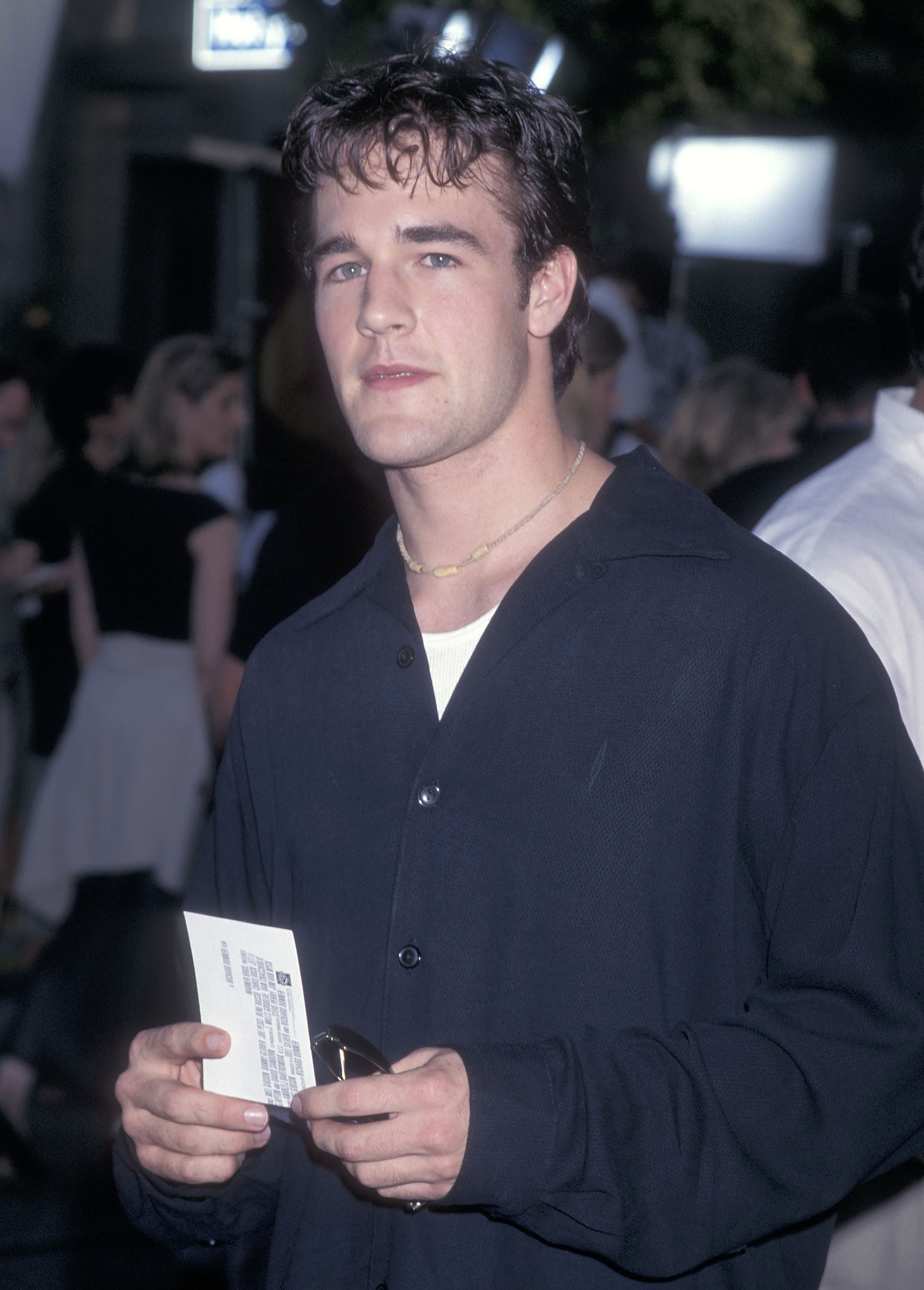 James Van Der Beek stands amid the premiere crowd, holding a ticket as soft lights illuminate the scene around him. His dark shirt and simple necklace give him a relaxed, understated style against the lively backdrop.