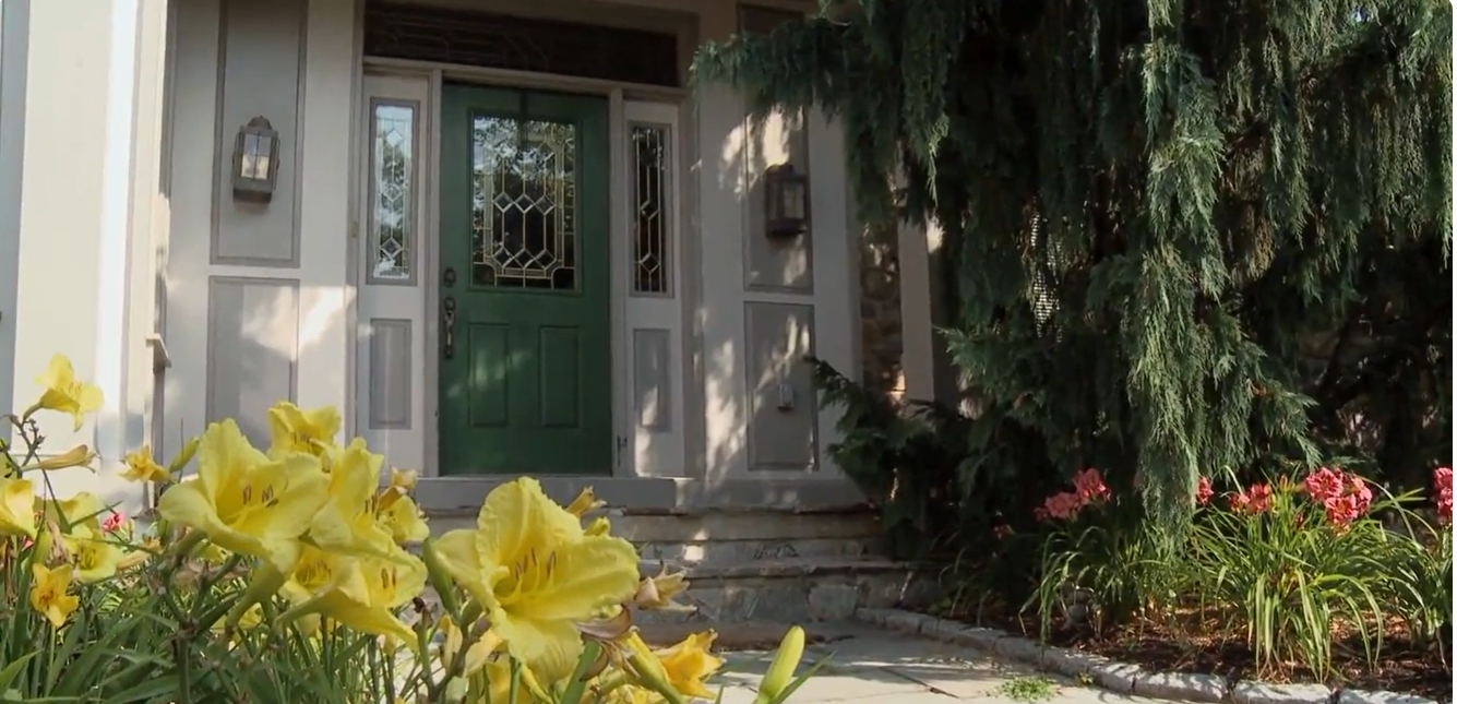 The front entrance of the Gosselin home features a dark green door with decorative glass panels, narrow sidelights, and traditional wall lanterns. Stone steps lead to the entrance, surrounded by yellow flowers, pink blooms, and a large evergreen tree. | Source: YouTube/TLC