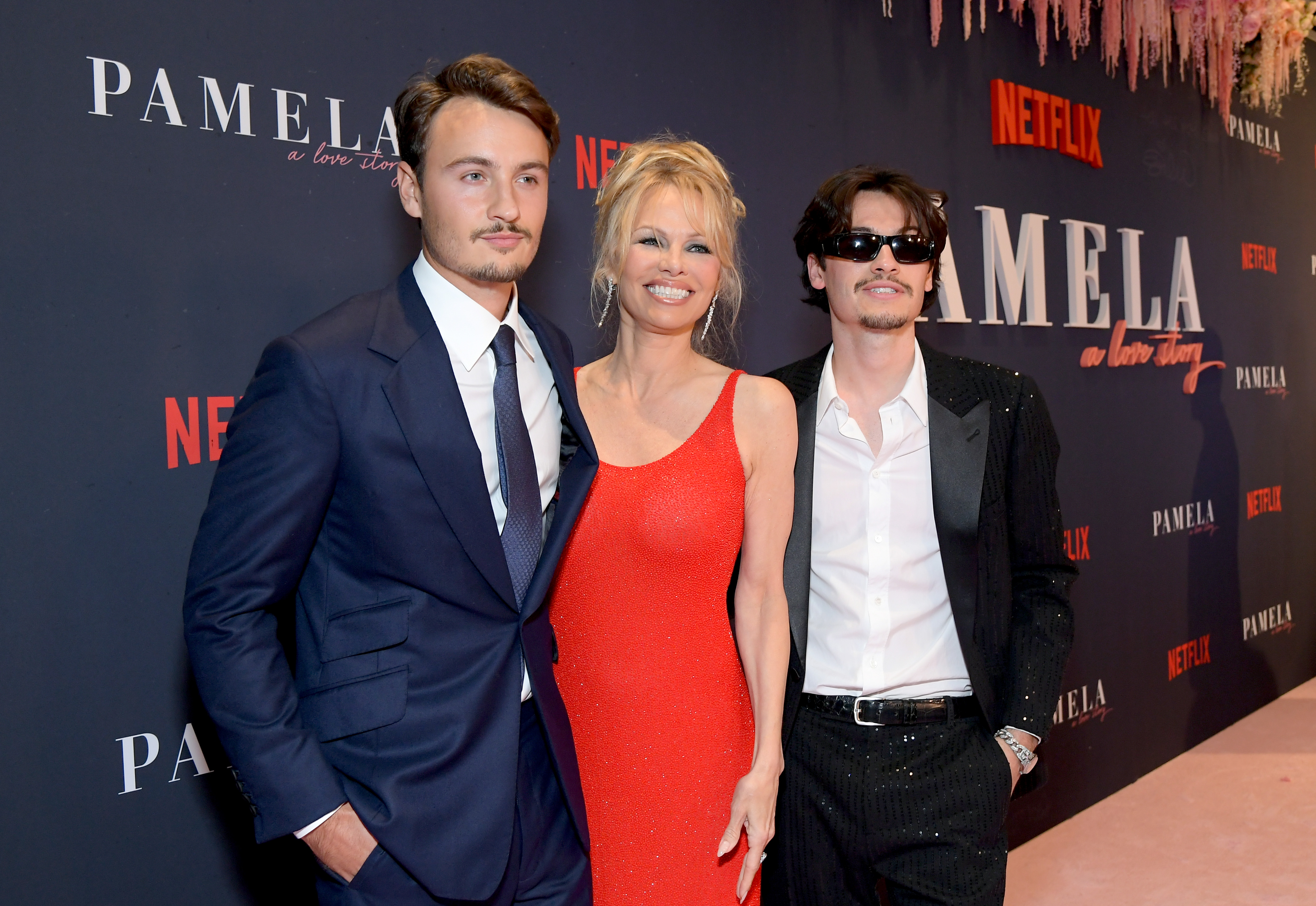 Brandon, Pamela, and Dylan attend the Los Angeles premiere of Netflix’s "Pamela," a love story at the Netflix Tudum Theater on January 30, 2023. Pamela wears a red, fitted gown while Brandon appears in a navy suit and Dylan in a black sequined jacket and sunglasses.