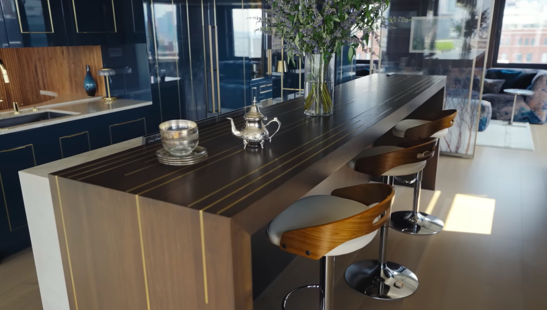 The kitchen counter is almost a piece of furniture in its own right, with delicate brass strips set into the dark surface. Curved wooden stools echo the soft, rounded shapes found throughout the rest of the apartment. | Source: YouTube/Architectural Digest