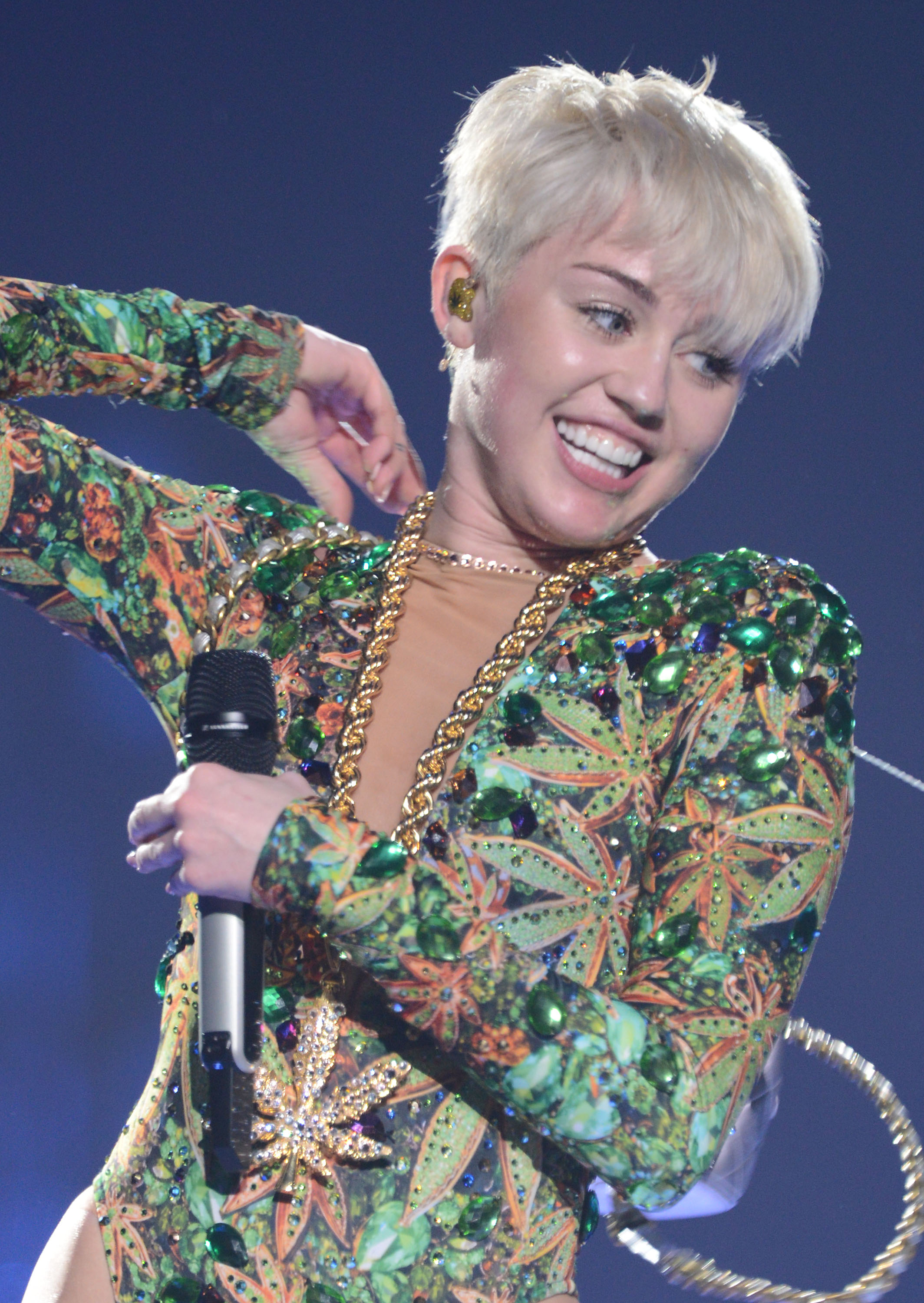 Miley Cyrus flashed a wide smile while performing with a microphone, wearing a striking green-and-orange embellished outfit, chunky gold chains, and a short platinum-blonde hairstyle. The stage shot captures a dramatic departure from her earlier Disney-era styling and the bold image she embraced as her music career evolved. | Source: Getty Images