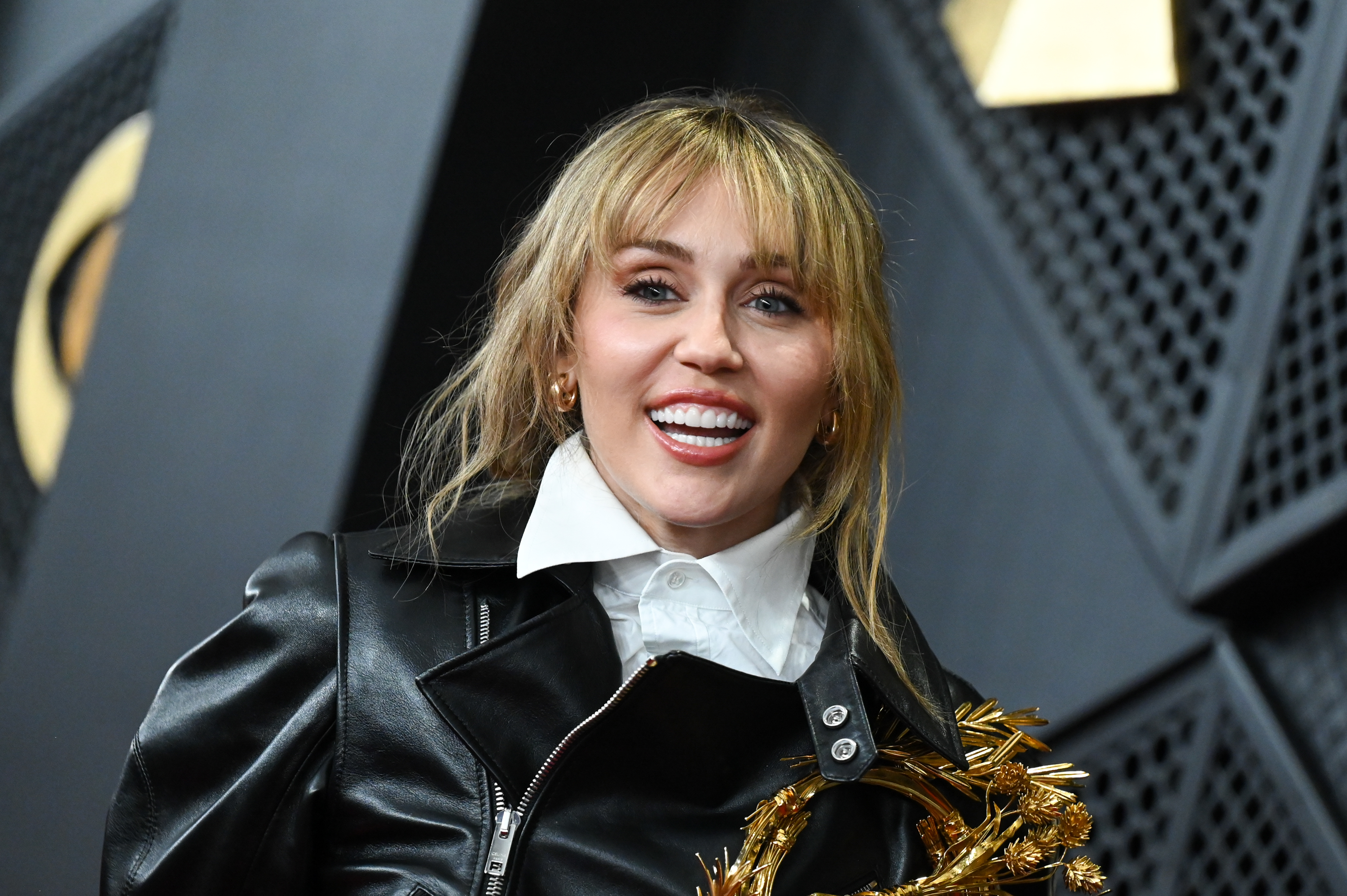 Miley Cyrus flashed a wide smile in a black leather jacket adorned with gold detailing. Her polished red-carpet appearance contrasts with the pressures she described in her conversation with Lewinsky, where she recalled growing up on a large farm before navigating a world where paparazzi, drones, and online commentary can make privacy difficult to maintain. | Source: Getty Images
