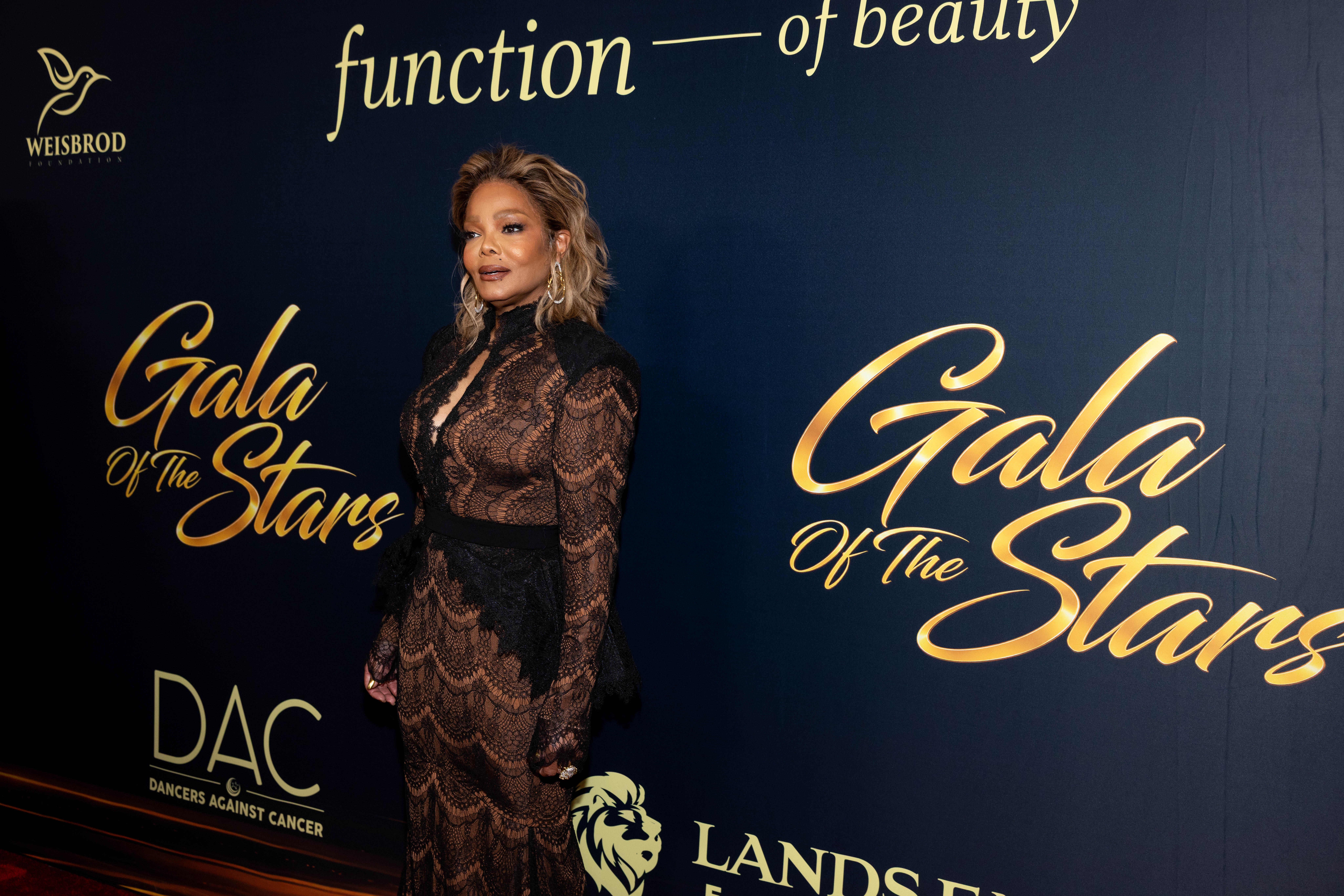 Janet Jackson poses in profile on the red carpet at the Gala of the Stars, displaying the fitted silhouette and intricate lace detailing of her sheer black gown against the event’s gold-lettered backdrop. | Source: Getty Images