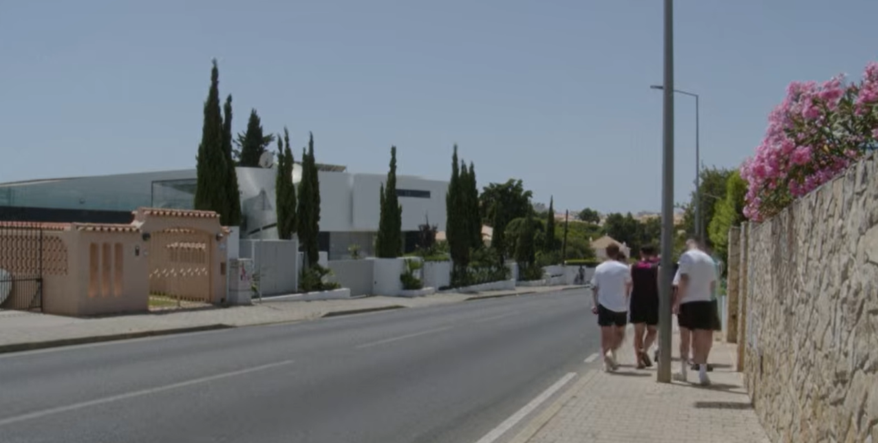Three pedestrians walk along a sunlit street near Bonnie Tyler’s modern Portugal home, its sleek white exterior and towering cypress trees creating a striking contrast with the quiet residential surroundings. | Source: YouTube/AFP News Agency