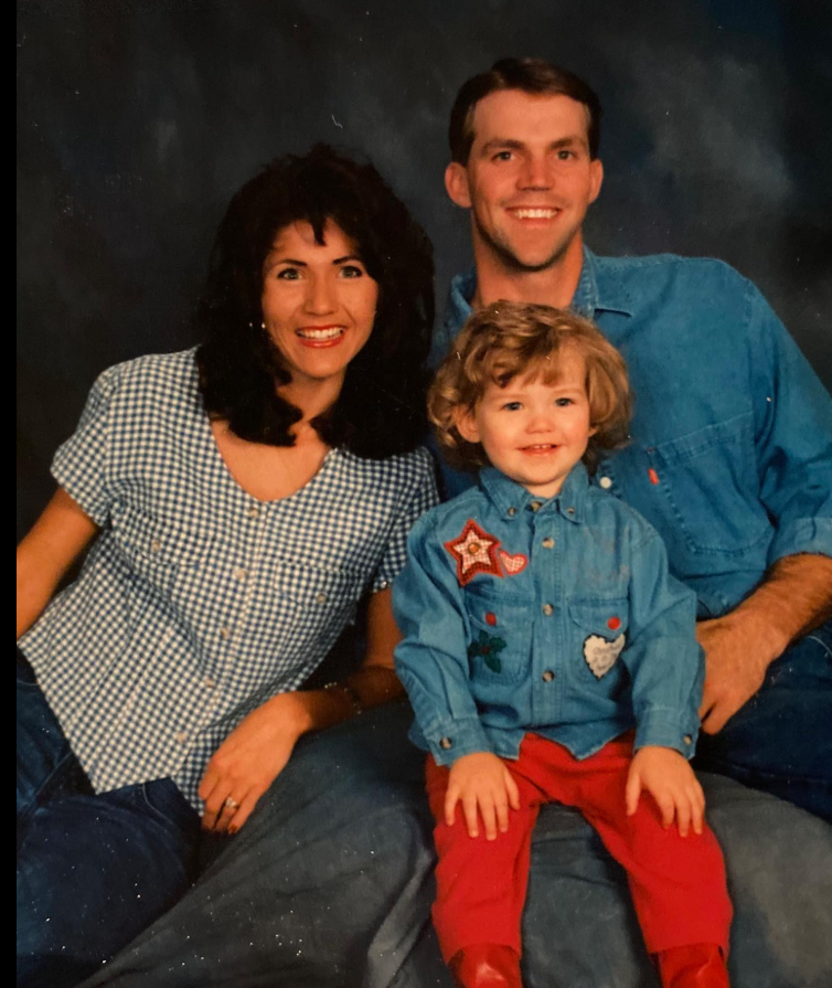 Bryon and Kristi Noem sit close, smiling with easy familiarity, as a young child in bright red pants and a denim shirt takes center stage—small, cheerful, and clearly the heart of the moment. | Source: Facebook/KristiNoem