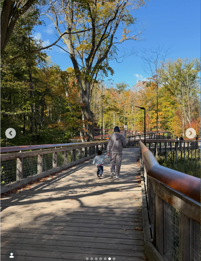 Bader Shammas walks hand in hand with his son, Luai, along a wooden boardwalk surrounded by trees displaying vibrant autumn colors. The pair are photographed from behind beneath a clear blue sky, with sunlight casting long shadows across the path. | Source: Instagram/lindsaylohan